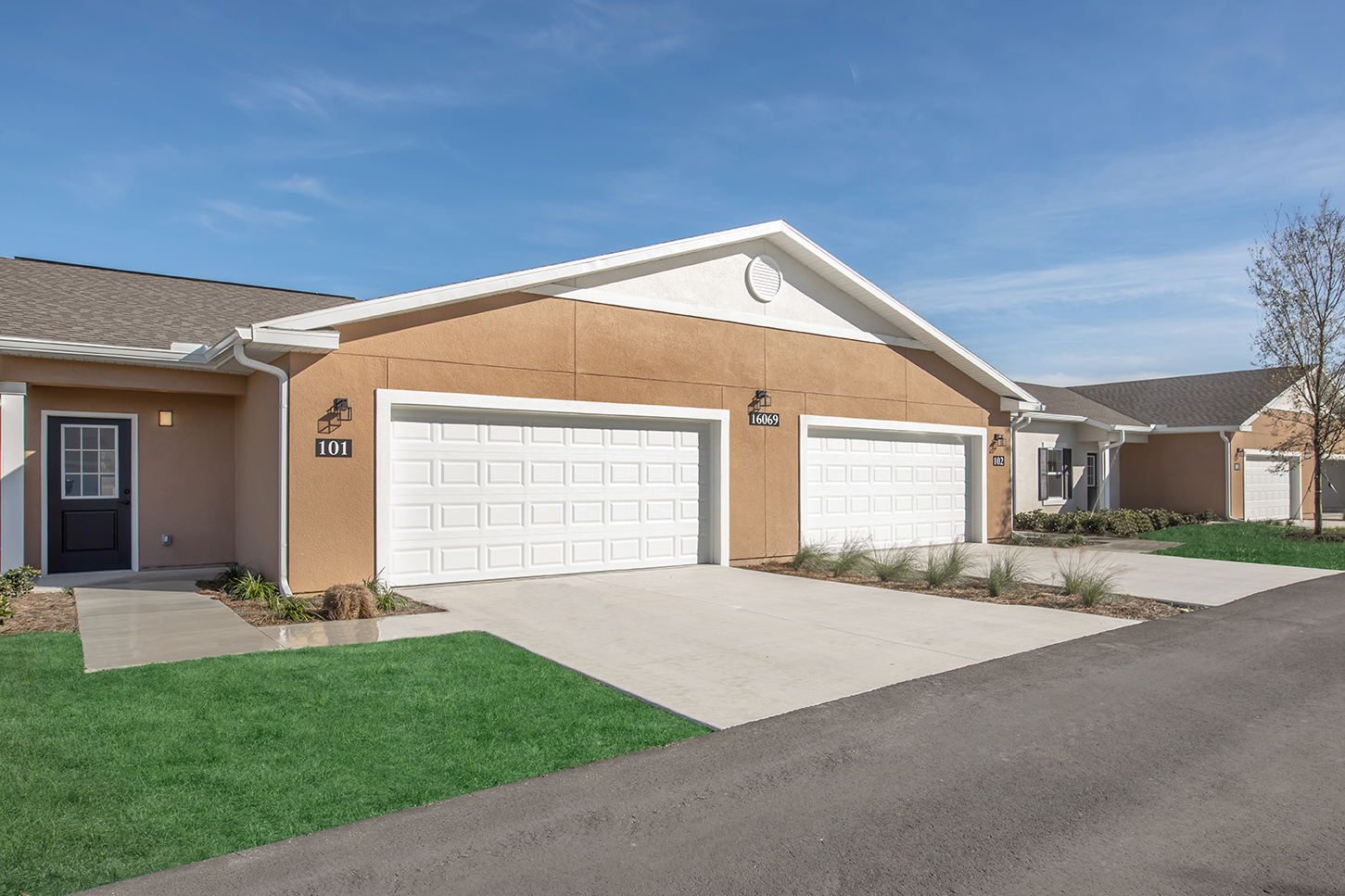 Two residential units with garages, featuring light brown exteriors and white garage doors, are situated side by side. The pavement leads to a grassy area, and the sky is clear with a few clouds, indicating a sunny day. The units are located in a suburban neighborhood.