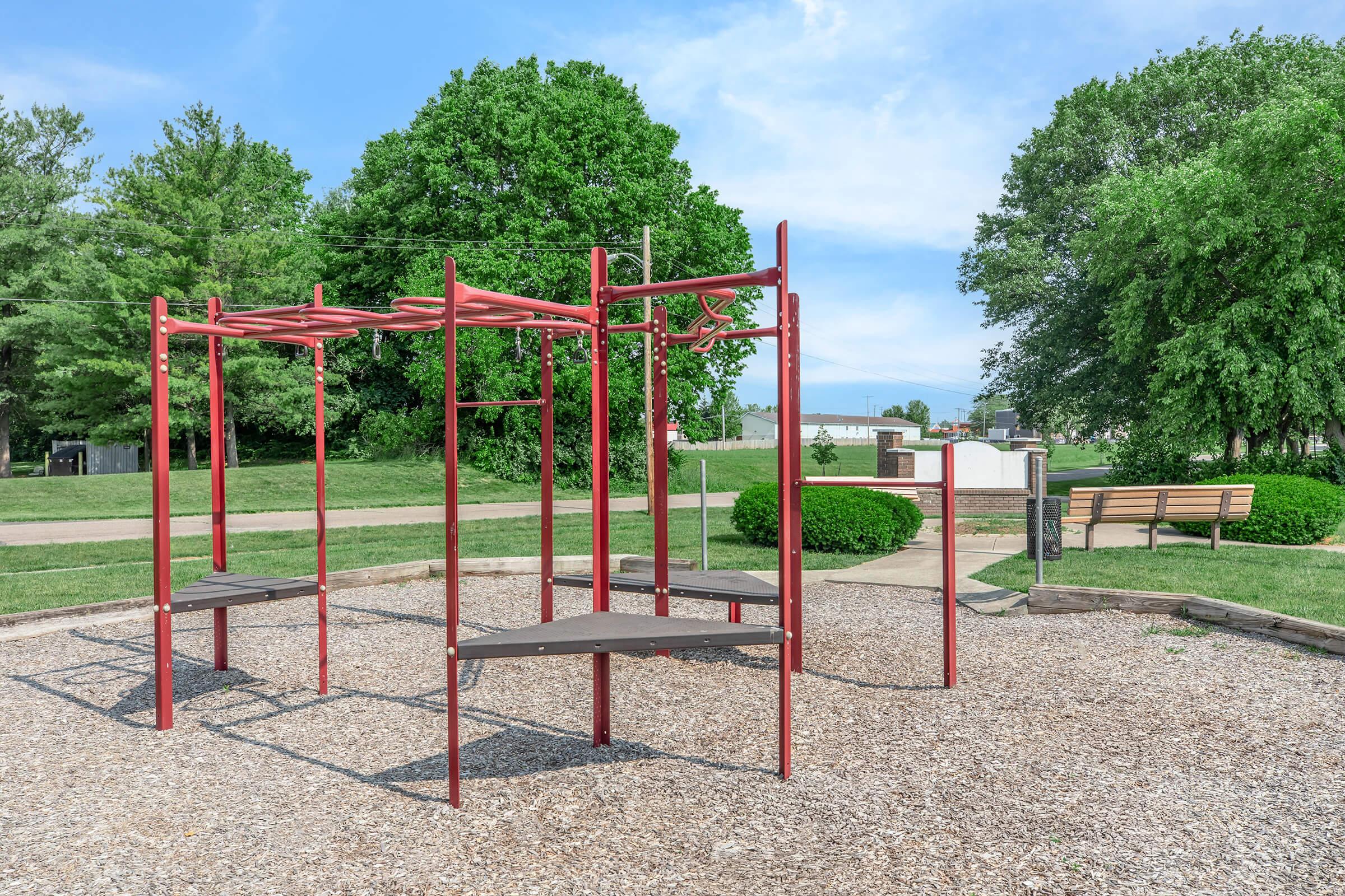 A playground featuring red climbing equipment with wooden platforms, surrounded by a gravel surface. In the background, there are green trees and a bench in a landscaped area, creating a serene outdoor environment. Clear blue sky enhances the bright scene.