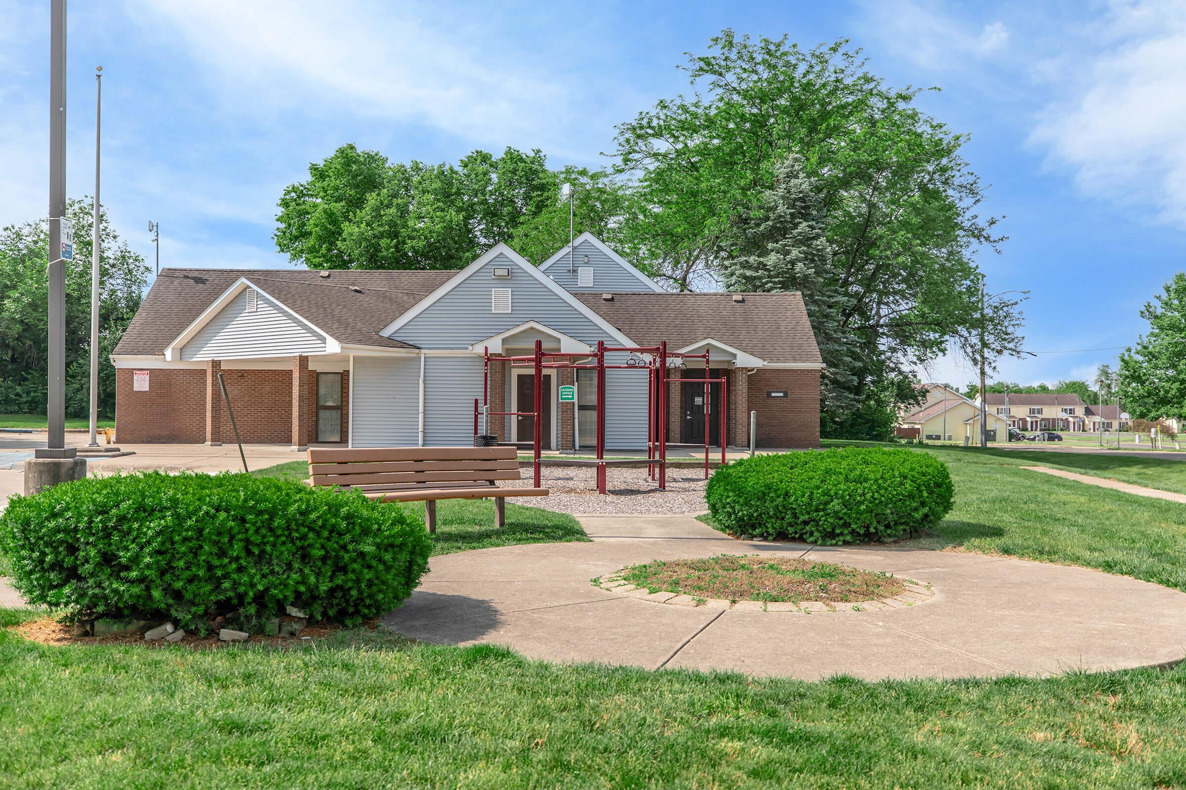 A small community center surrounded by green grass and trees. In the foreground, there is a circular garden feature and a wooden bench. To the side, there is a playground structure. The building has a sloped roof and multiple entrances, with a flagpole visible in the background.