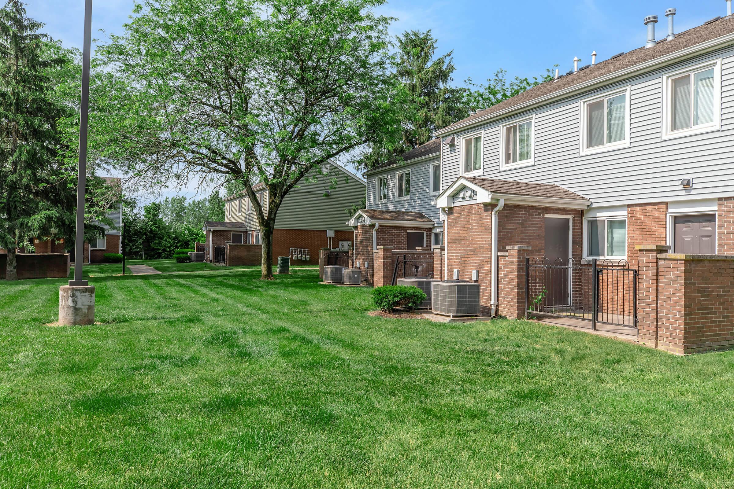 Row of residential buildings with brick facades, located in a landscaped area featuring well-maintained green grass and trees. Air conditioning units are visible outside some units, and pathways lead between the buildings. Clear blue sky overhead enhances the peaceful suburban setting.