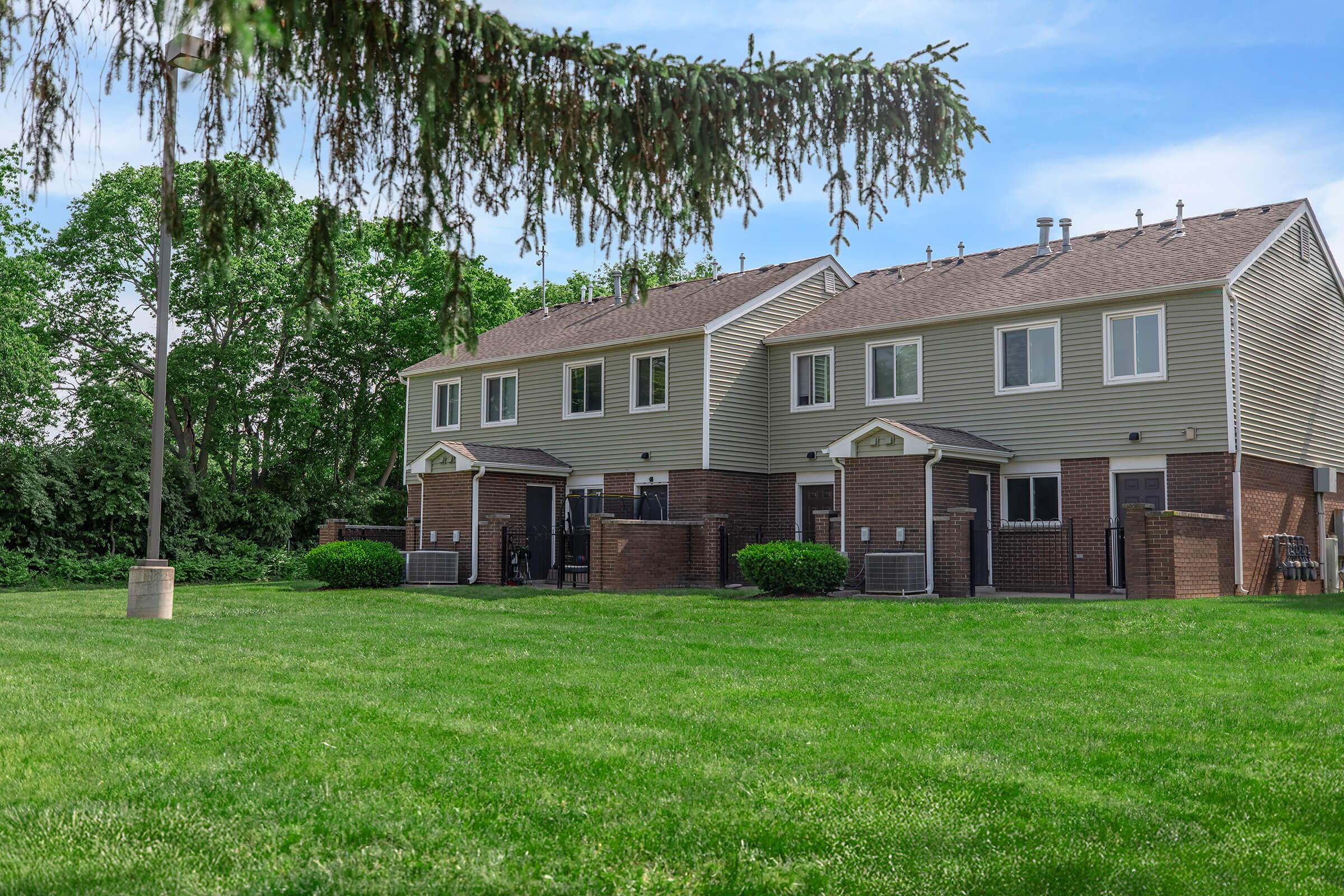 A row of two-story townhouses with brick and siding exteriors, situated on a green lawn. Trees line the background, and each townhouse has a small patio area with air conditioning units visible. The scene conveys a residential setting with an emphasis on greenery and outdoor space.