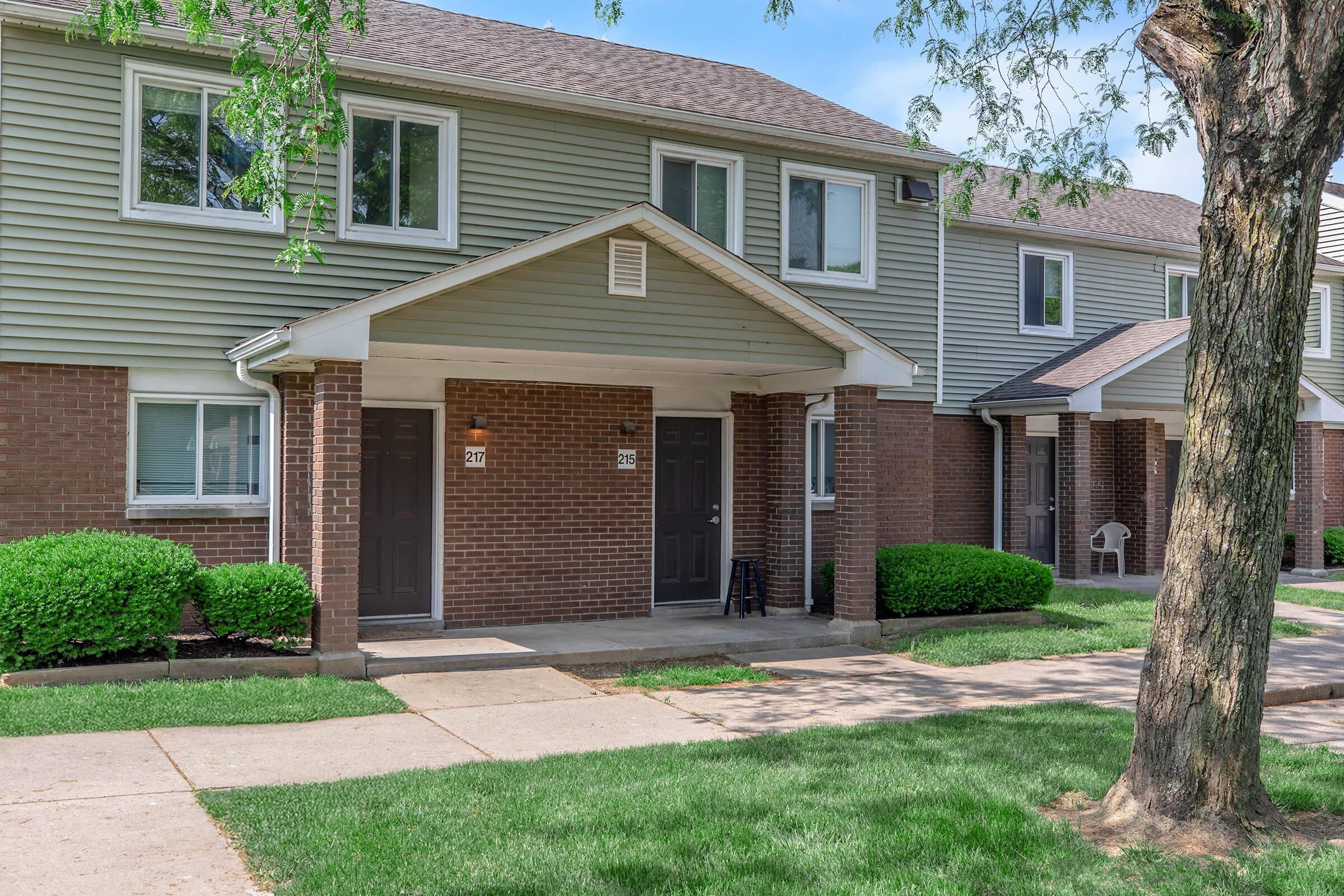 Two-story residential building with multiple units. The façade features light green siding and brick accents. Each entry has a dark door, and small shrubs are neatly trimmed in front. A tree is visible on the right side, and the sidewalk leads up to the doors, suggesting a well-maintained neighborhood.