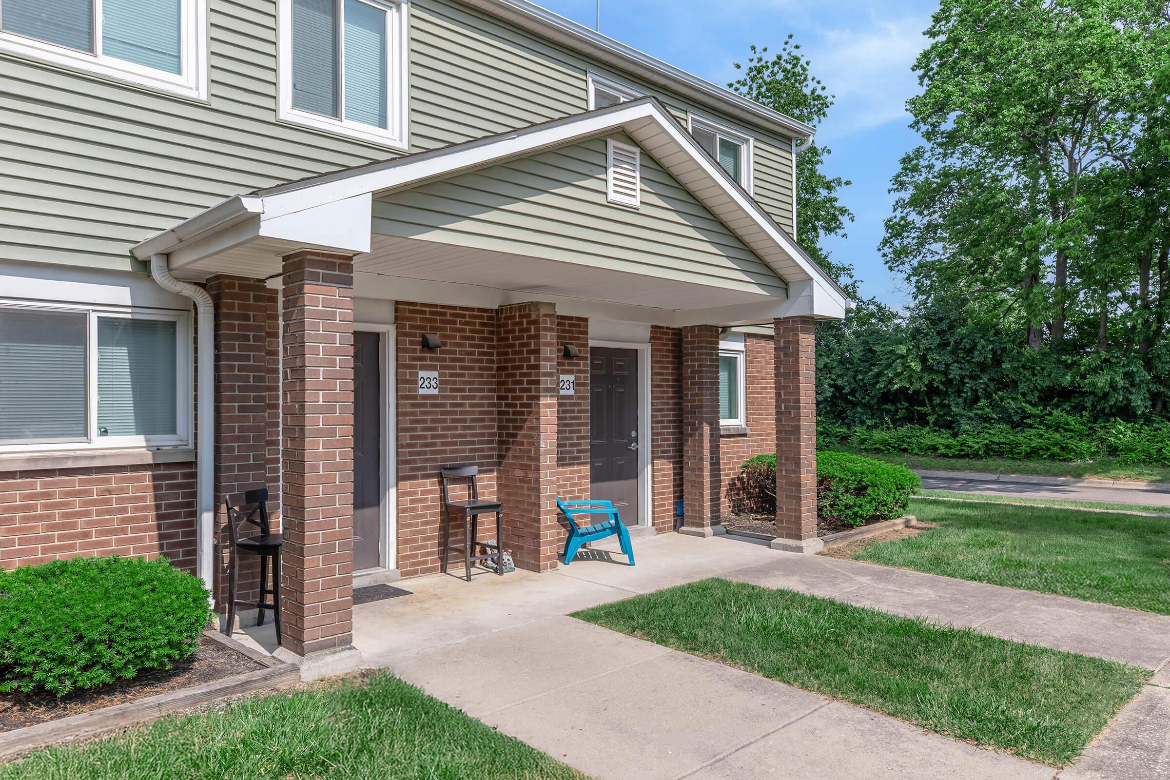 A two-story apartment building with a light green exterior and brick accents. Two entrance doors are visible, along with a small porch area featuring a chair and a blue patio chair. The surroundings include neatly trimmed grass and shrubs, under a clear blue sky.