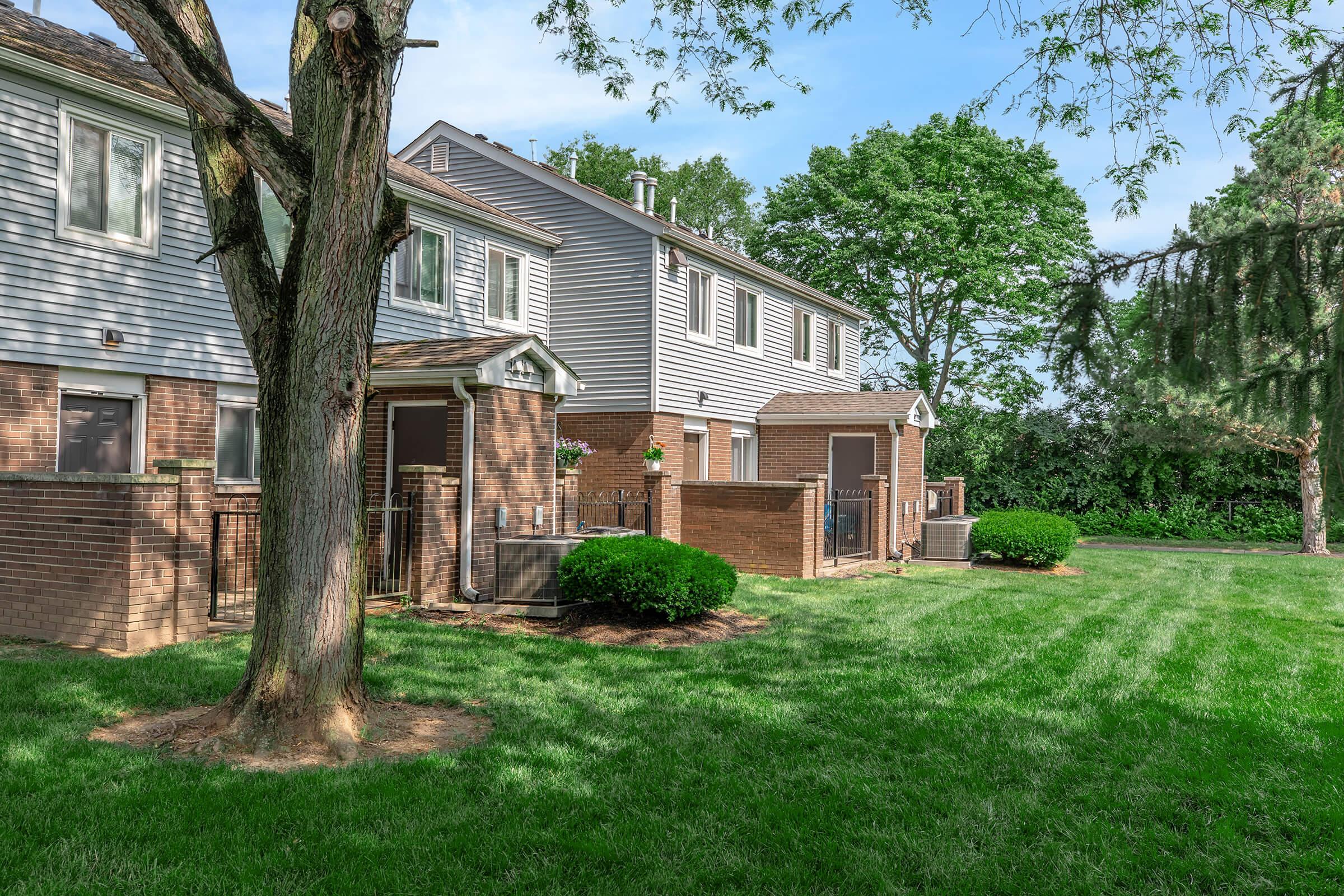 Row of townhouses in a residential area, featuring a well-manicured lawn with green grass, shrubs, and trees. The buildings have gray siding and brick accents, and there is a fence surrounding the backyards. The scene is well-lit with blue sky and soft clouds in the background.