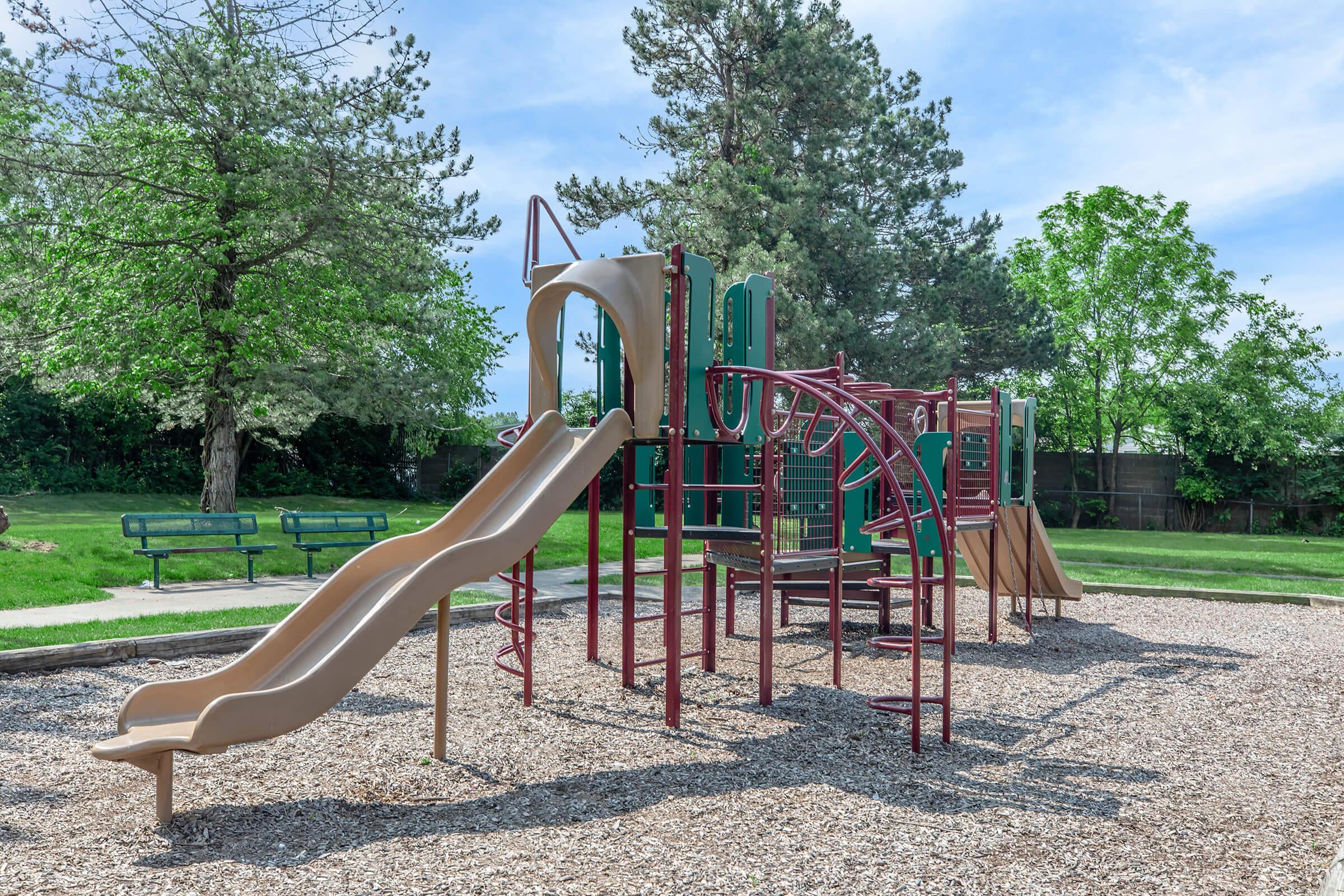 A playground featuring a beige slide and various climbing structures, surrounded by gravel and green grass. In the background, there are benches and trees under a clear blue sky.