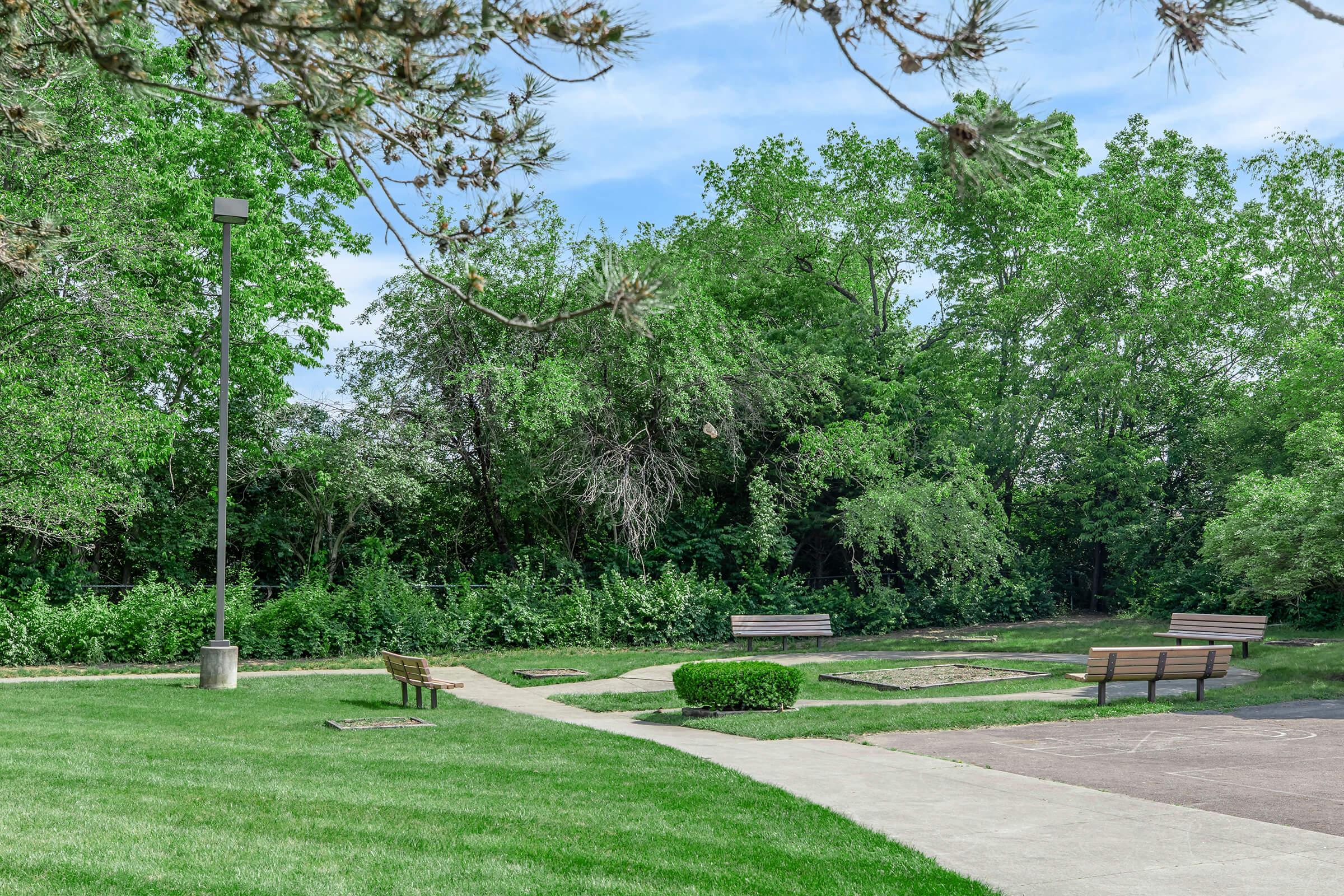 A landscaped park area featuring green grass and trees. There are benches arranged along paths that curve through the space. The scene includes a light post, with a backdrop of lush foliage, creating a serene and inviting outdoor environment. Ideal for relaxation and enjoying nature.