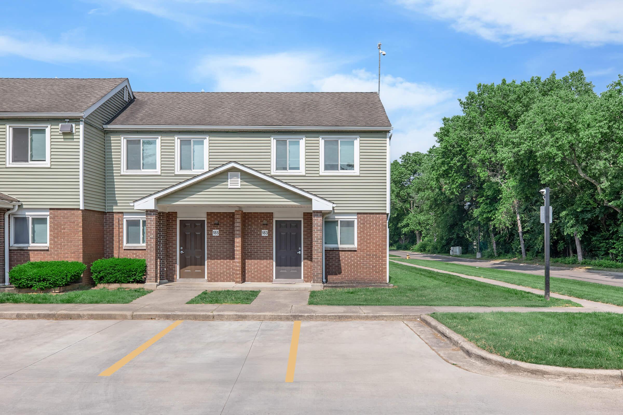 A two-story residential building with a beige exterior and brown brick accents, featuring front door entrances with small porches. Well-maintained green grass and shrubs surround the building, and a paved parking area is visible in the foreground. A tree-lined road is seen in the background under a clear blue sky.