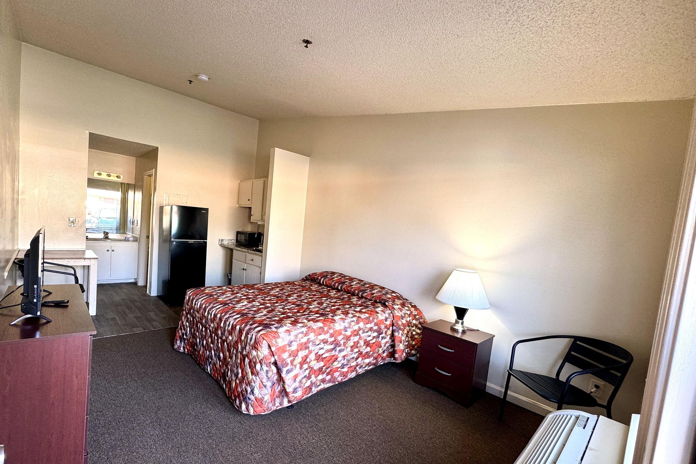 A cozy hotel room featuring a queen-sized bed with a patterned comforter, a nightstand with a lamp, a small table with a chair, a flat-screen TV, and a kitchenette area. The walls are light-colored, and there is a window providing natural light. The floor is carpeted, and the overall atmosphere is inviting and simple.