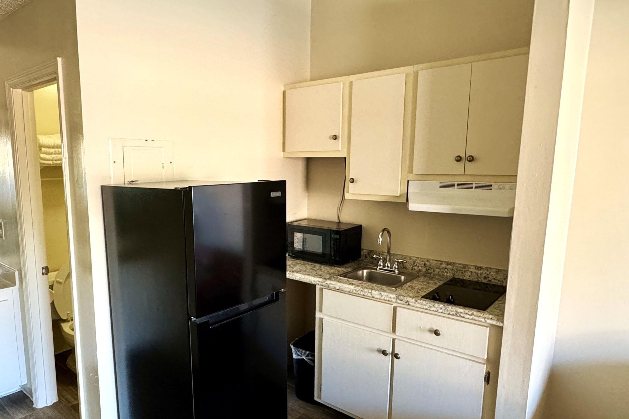 A compact kitchen featuring a black refrigerator, a microwave, a sink, and a stovetop. The cabinets above and below the countertop provide storage, while the room has light-colored walls and a hardwood floor. A glimpse of a restroom can be seen in the background.