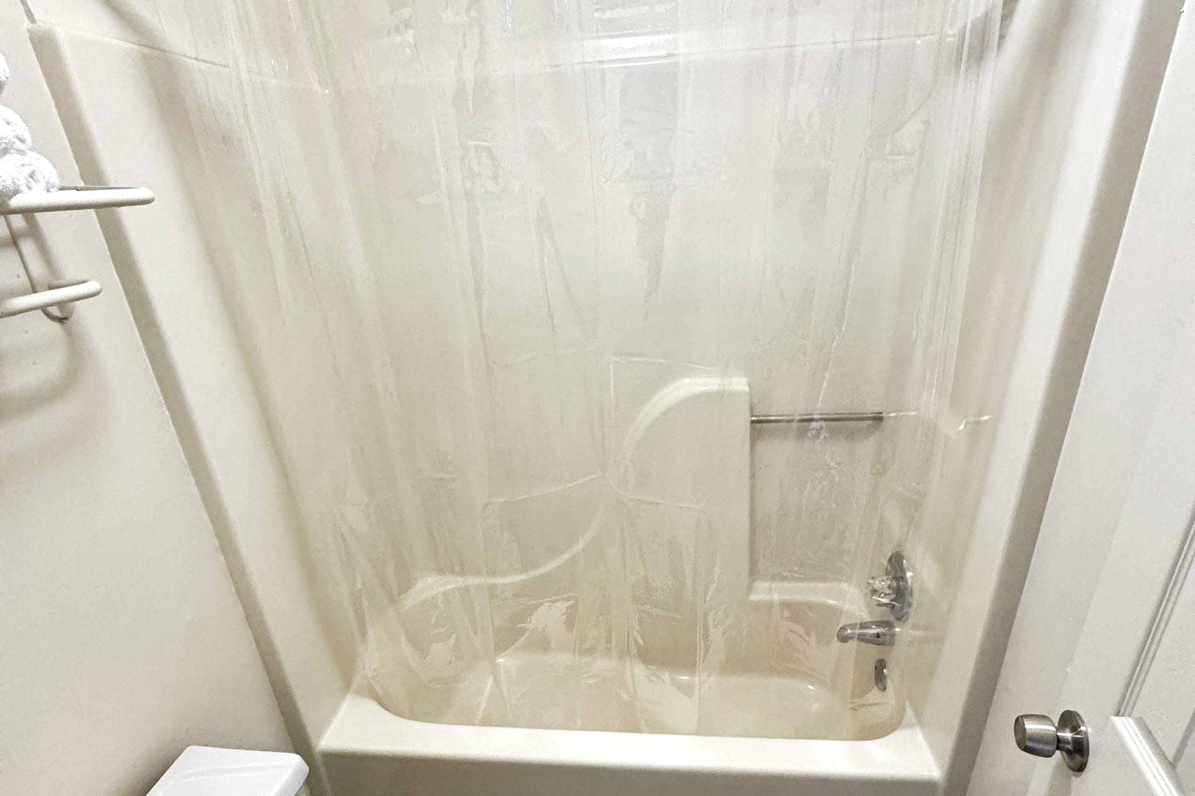A bathroom shower space with a clear plastic curtain partially drawn. The tub area has a light-colored wall and a faucet. A small white shelf holds neatly folded towels on the left. A toilet is visible at the bottom left, and there's a door handle on the right side of the image.