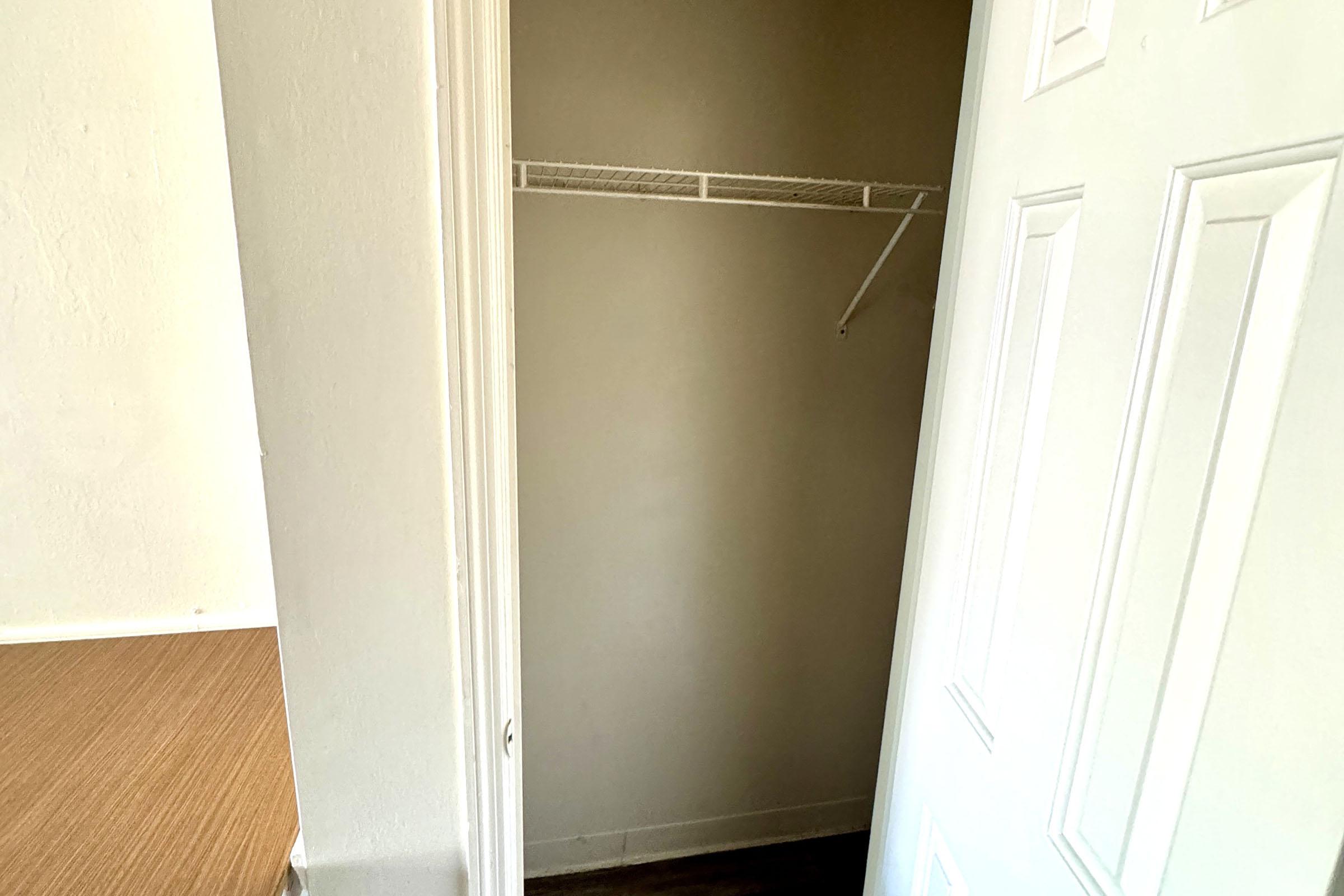 A view of a small, empty closet with a white wire shelf and a light beige wall. The closet door is slightly open, revealing the interior space, which has a clean, minimalistic look. Adjacent to the closet is a wooden floor and a wall that appears to be painted in a light color.