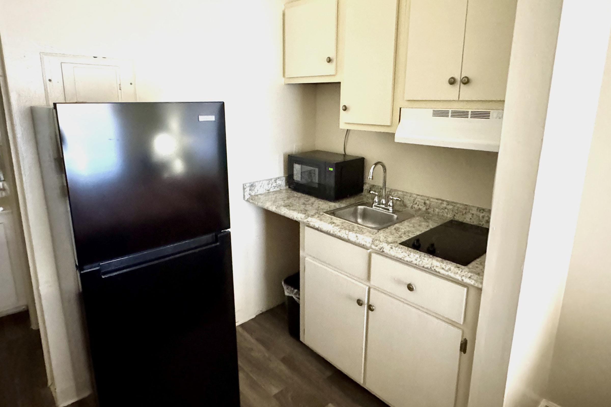 A small kitchen featuring a black refrigerator, a stainless steel sink, a microwave, and a countertop with a stovetop. The cabinets are light-colored, and the flooring is wooden. The space is tidy and functional, designed for basic cooking and food storage.