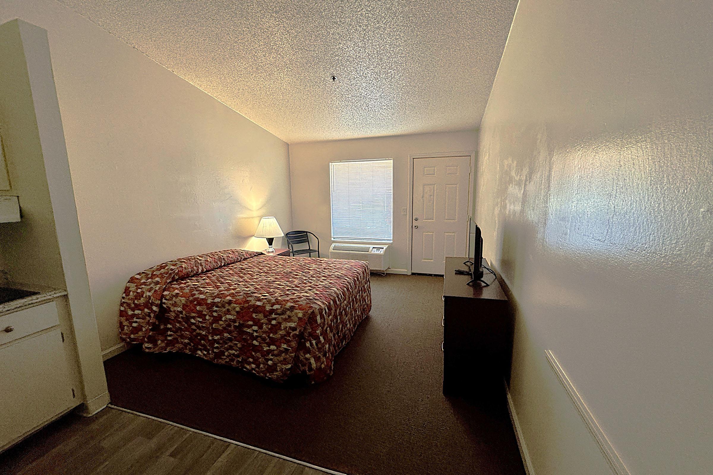 A simple hotel room featuring a bed with a patterned cover, a small nightstand, and a television. There is a window with natural light coming in, and a chair positioned nearby. The walls are painted white, and a kitchenette area is visible in the background. The floor is covered with carpet.