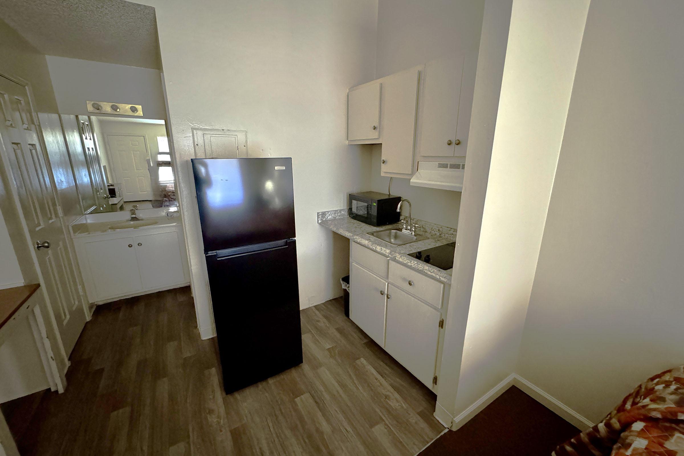 A small, compact kitchen area featuring a black refrigerator, a microwave on the countertop, and a sink. White cabinets and countertops add to the clean look, while laminate flooring completes the modern design. A small table space is visible in the corner, contributing to the cozy atmosphere.
