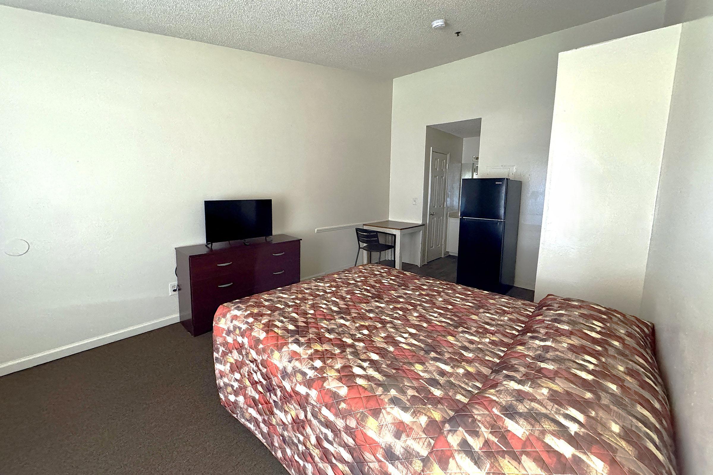 A simple hotel room featuring a patterned bedspread on a double bed, a small desk with a chair, a wall-mounted TV, and a refrigerator. The room has light-colored walls and carpet, creating a bright and airy atmosphere.