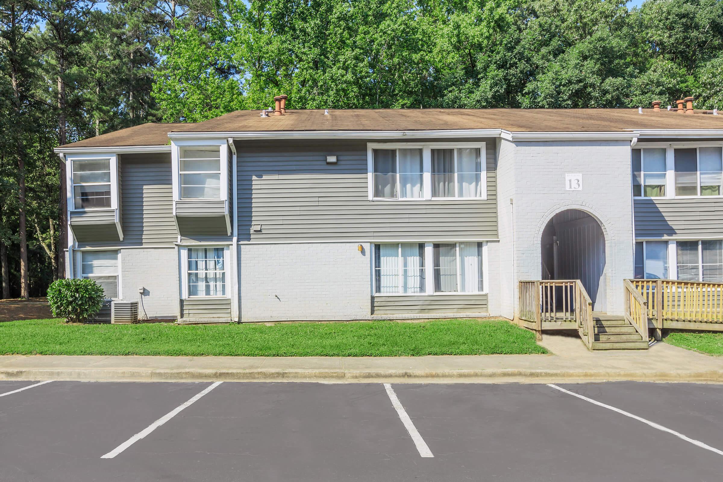 Exterior view of a two-story apartment building featuring multiple windows, a walkway leading to an entrance, and green lawn area. The building is painted in grey tones, with a light-colored brick section. Several parking spaces are visible in front of the building.