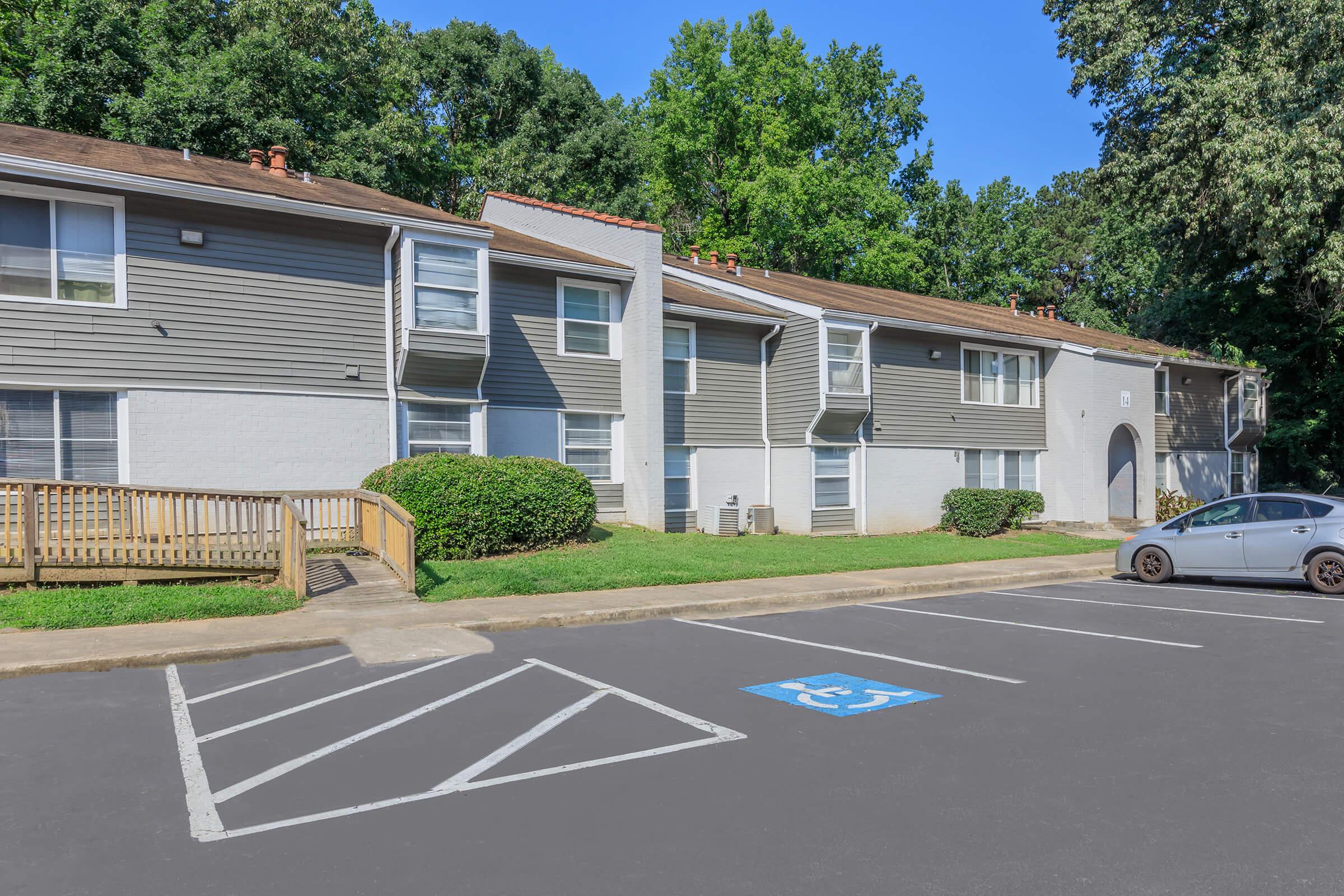 Two-story apartment building with gray siding and white accents, surrounded by trees. A wooden railing leads to the entrance, and a designated wheelchair parking space is visible in the foreground. Sunlight illuminates the scene, creating a welcoming atmosphere.
