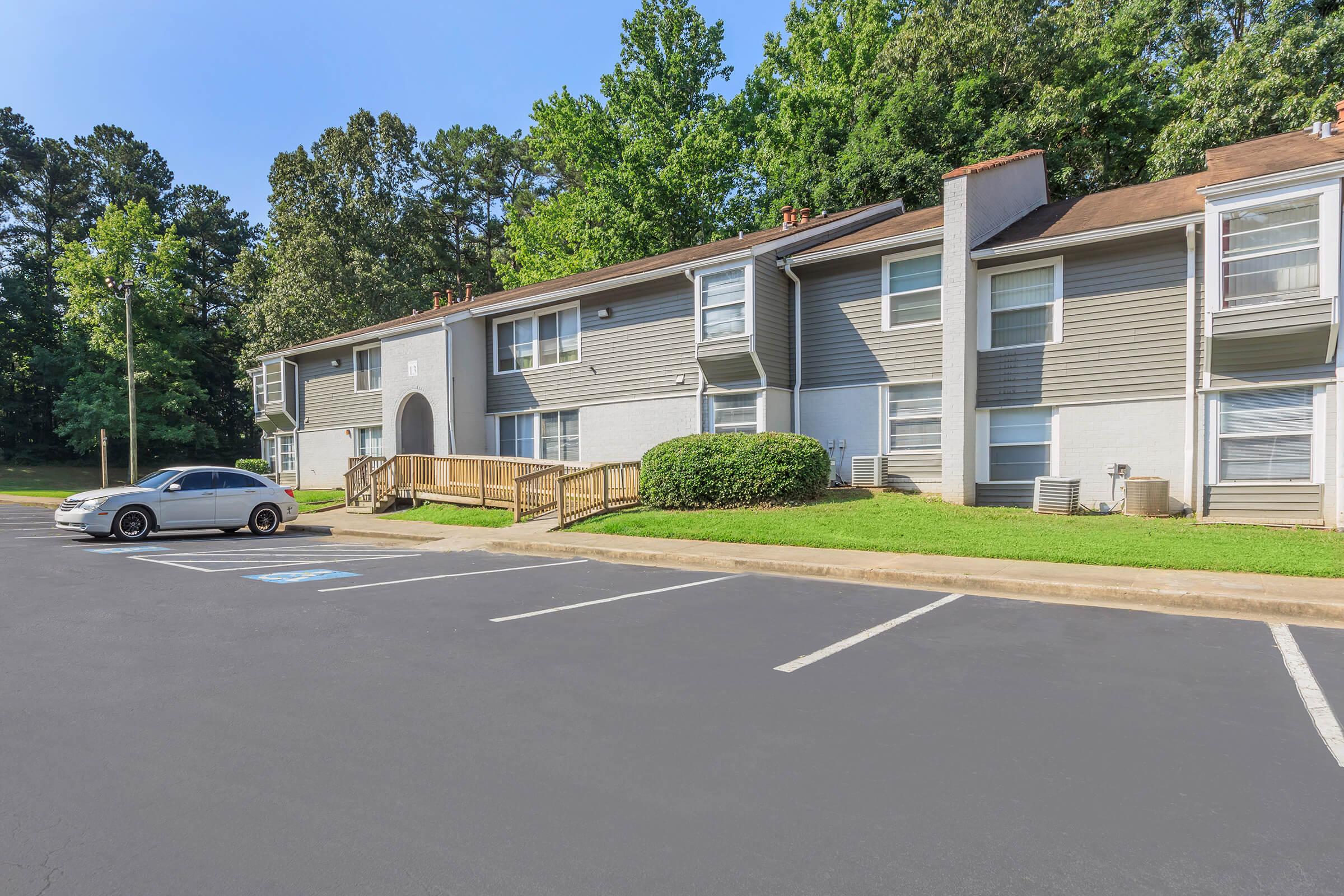 A multi-unit apartment building with a landscaped area, featuring a wooden ramp leading to the entrance. Several parked cars are visible in the parking lot, surrounded by trees and greenery in the background on a clear day.