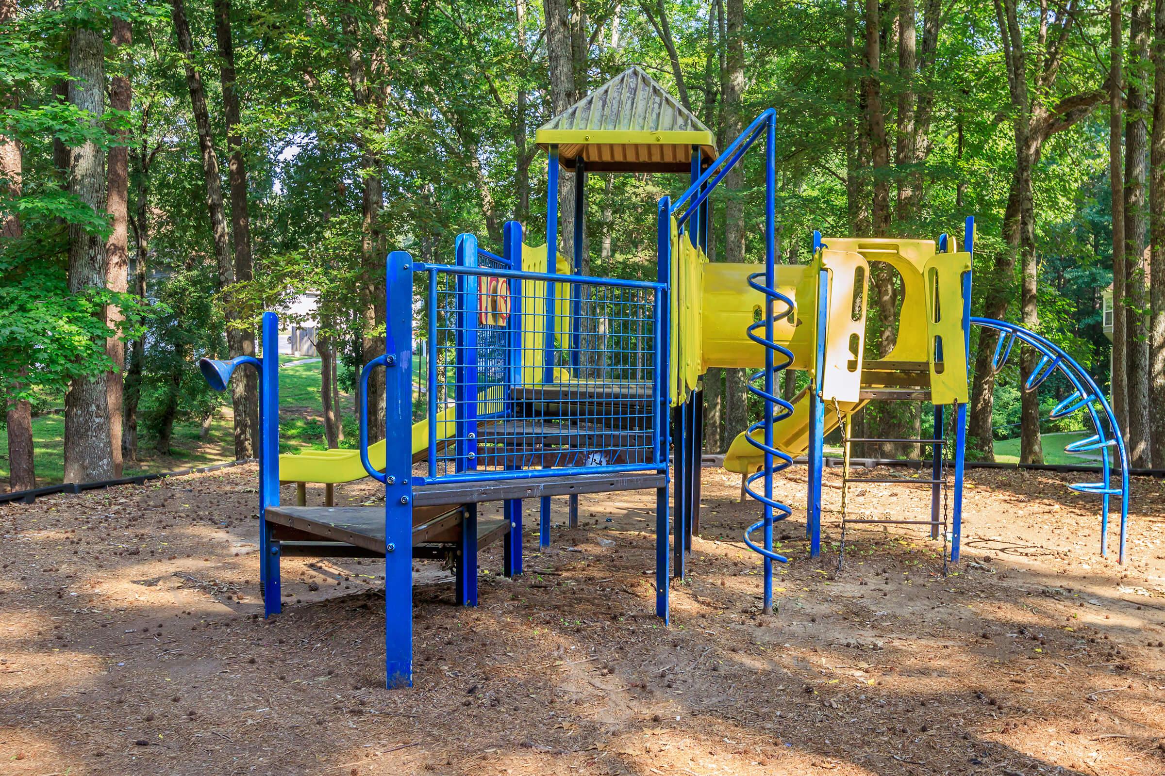 Colorful playground equipment in a wooded area, featuring a slide, climbing structures, and safety railings. Soft ground cover made of wood chips surrounds the play area, with lush green trees providing shade in the background.
