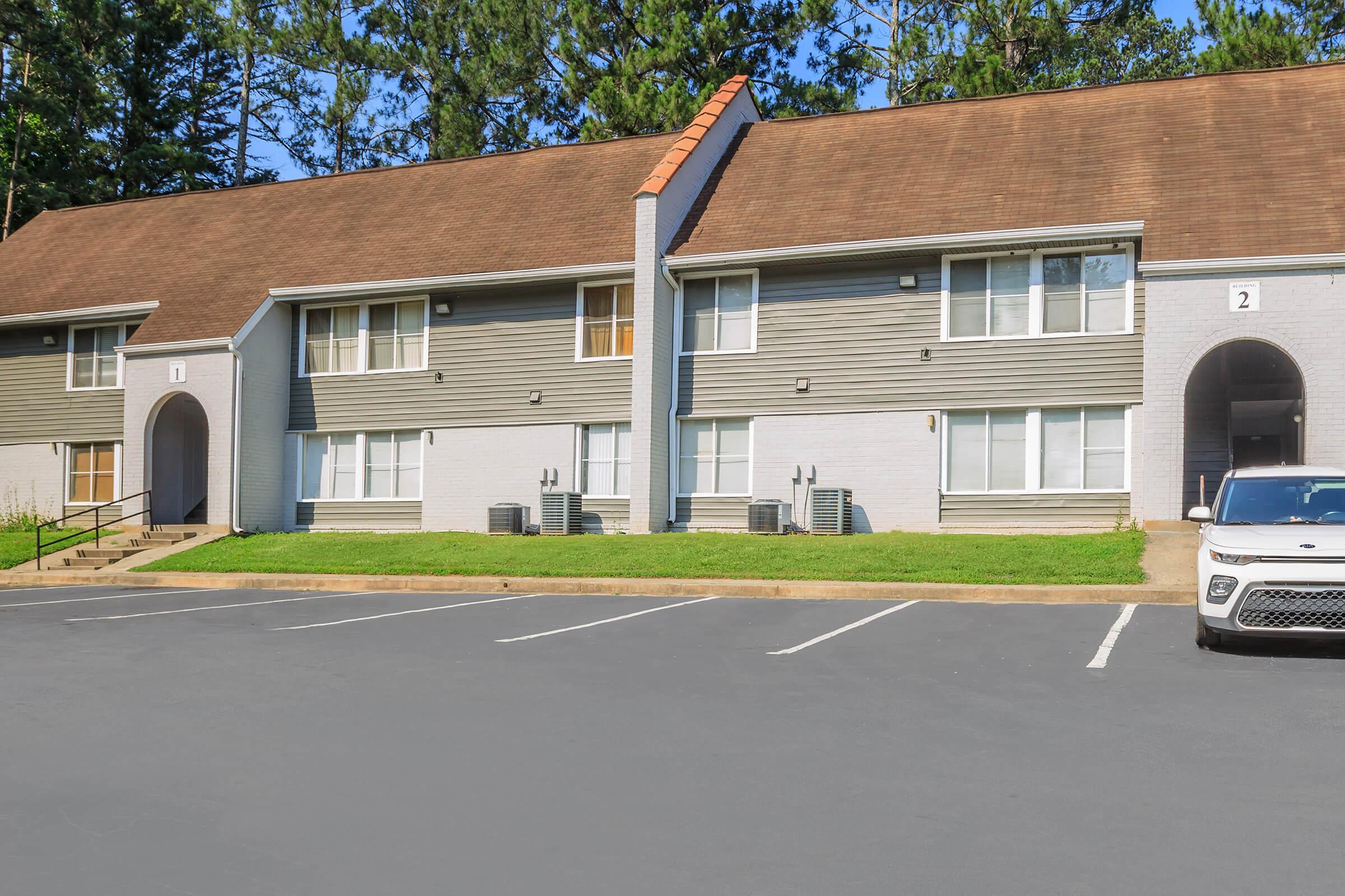 Two-story apartment buildings with a gray and beige exterior. The first building has two units with windows and air conditioning units visible. A white car is parked in front of the second building. The surrounding area includes a well-maintained lawn and trees in the background.