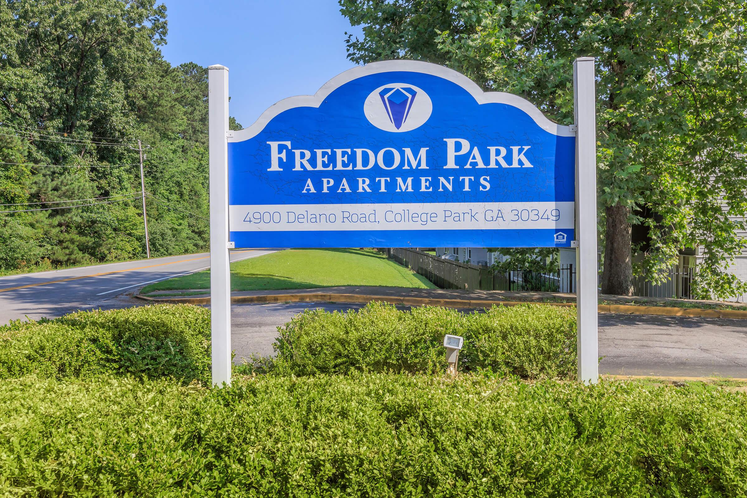 Sign for Freedom Park Apartments, featuring a blue background with white text. Includes address: 4900 Delano Road, College Park, CA 30349. Surrounded by greenery and trees in the background with a road visible nearby.