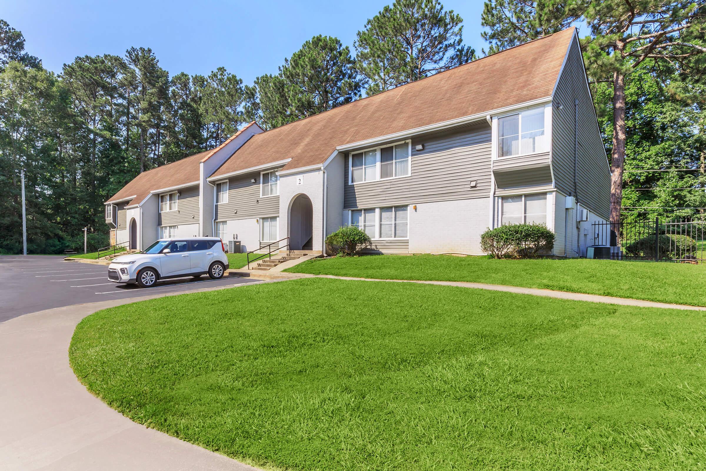 A two-story residential building with grey siding and a brown roof, surrounded by green grass and trees. A white car is parked in the driveway, and there are clear pathways leading to the entrance. The scene is sunny and well-maintained, conveying a peaceful suburban environment.