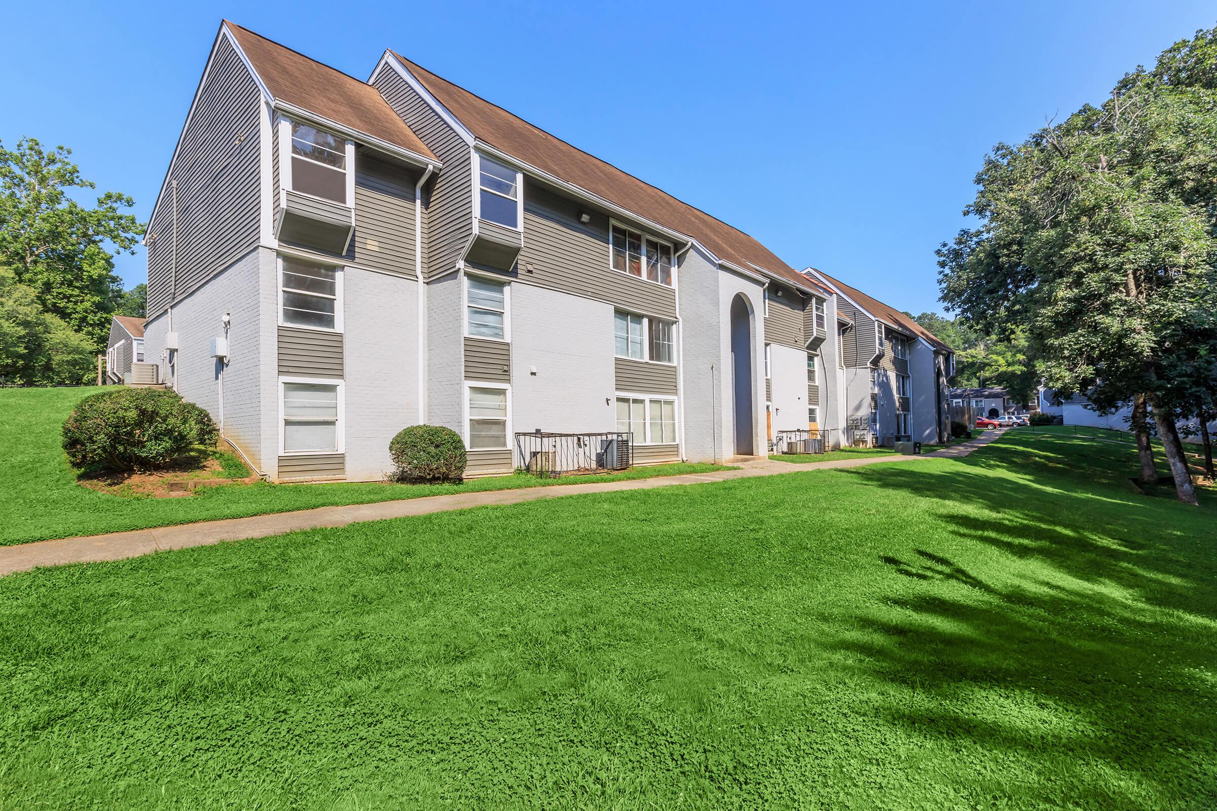 A row of multi-story apartment buildings with gray siding and brown roofs. The buildings are surrounded by well-maintained grassy areas and trees. The sky is clear and blue, creating a bright and inviting atmosphere.