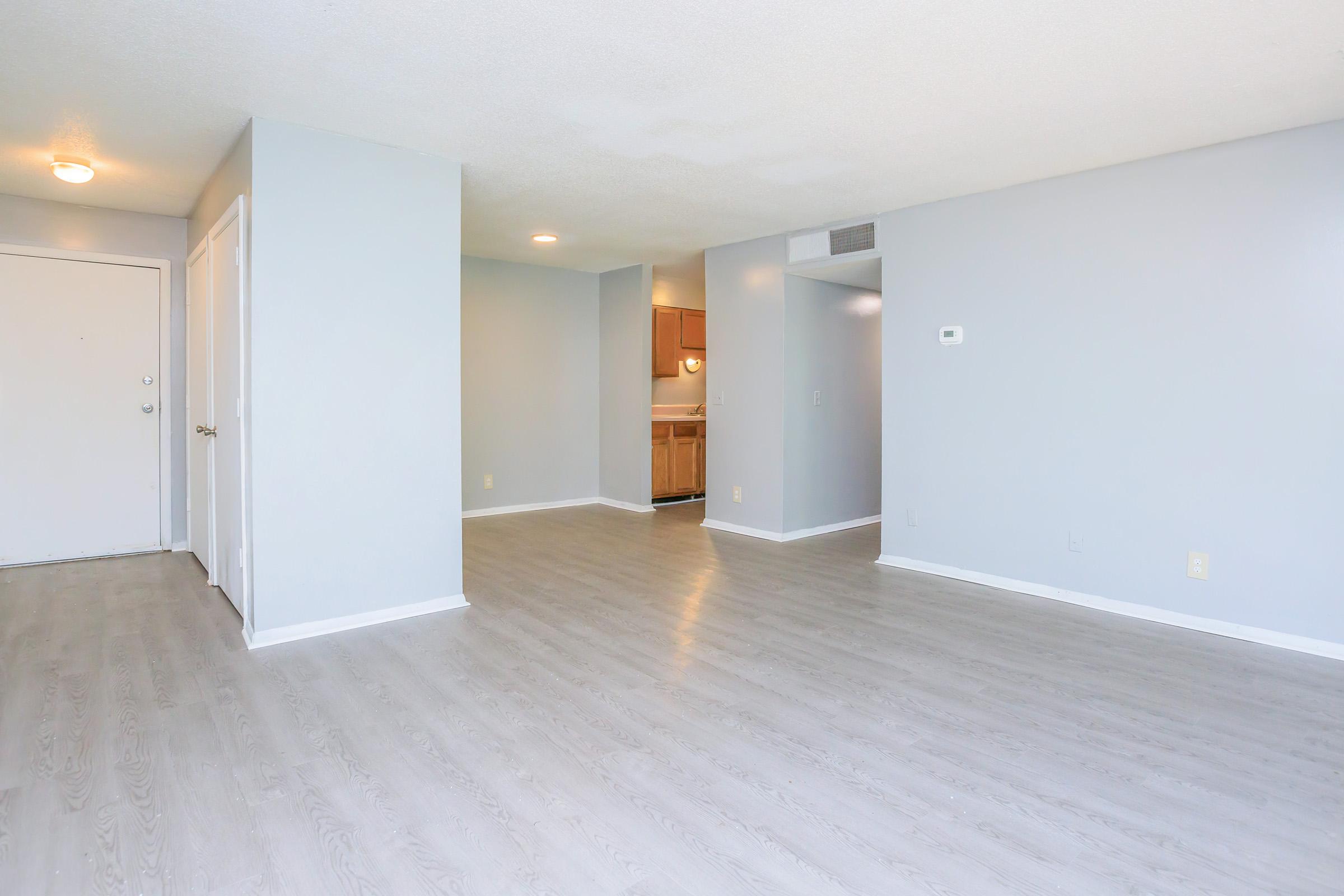 A spacious, empty room with light gray walls and laminate flooring. The entrance door is on the left, with a door leading to another room nearby. A kitchen area is visible in the background, featuring wooden cabinets. Natural light brightens the space, creating an inviting atmosphere.