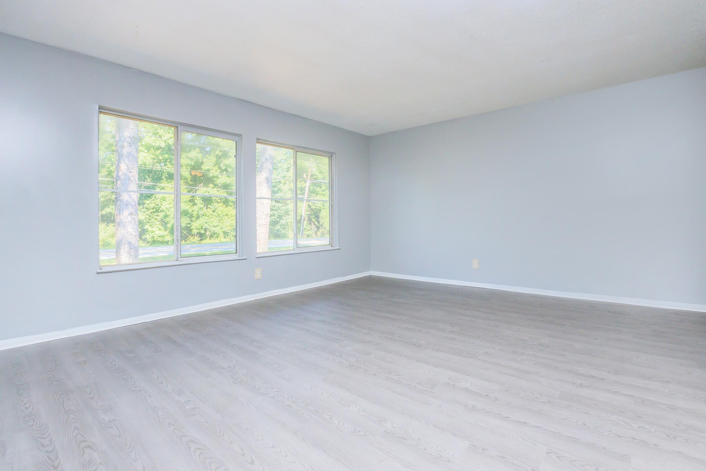 Spacious, empty room with light gray walls and large windows letting in natural light. The flooring is light-colored, enhancing the airy feel of the space. Green trees are visible outside through the windows, contributing to a serene atmosphere.