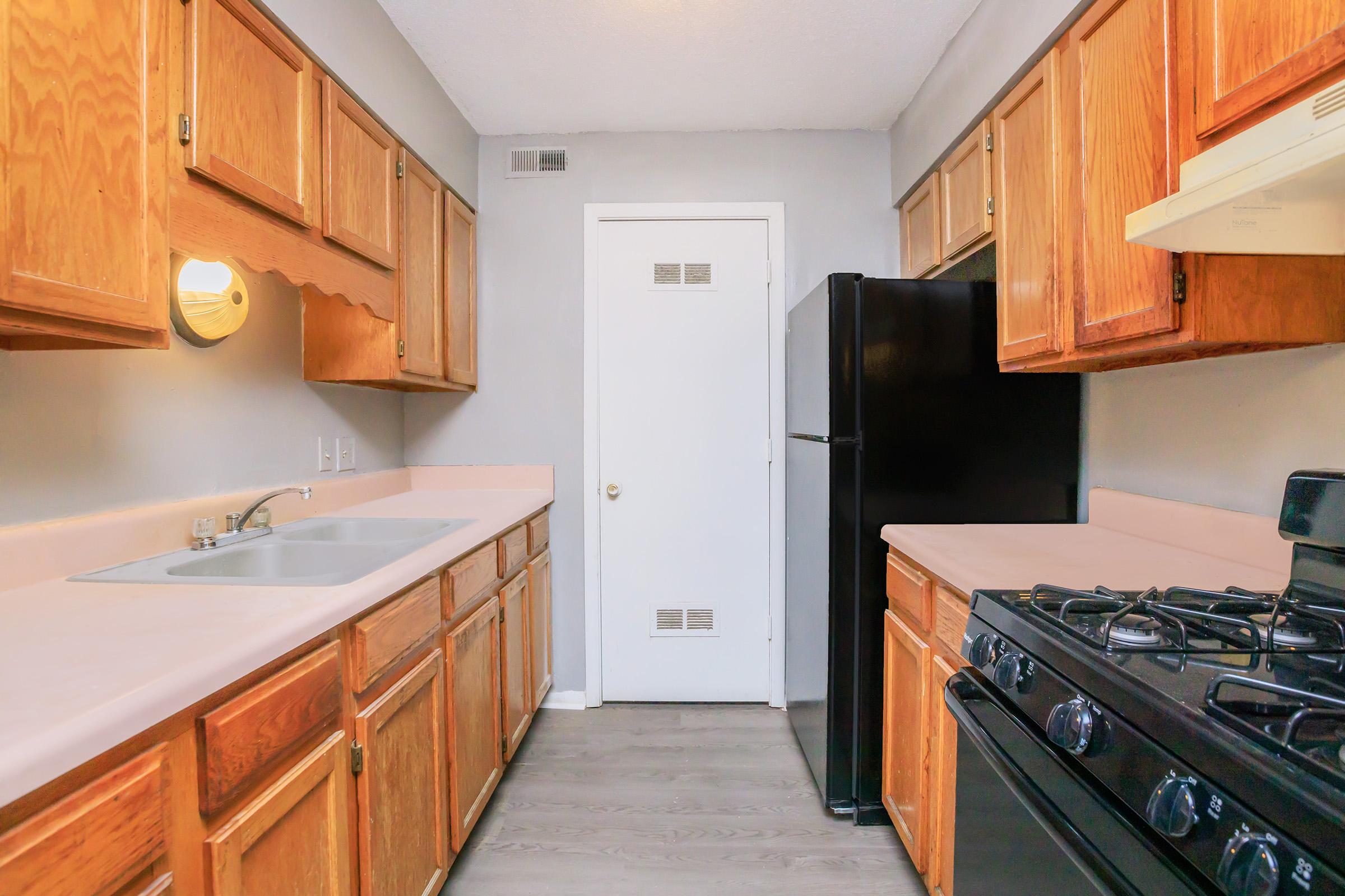 A narrow kitchen featuring wooden cabinets, a pink countertop, stainless steel sink, black gas stove, and a black refrigerator. The walls are painted light gray, and there's a closed door leading to another room. Bright lighting illuminates the space, enhancing its clean and modern look.