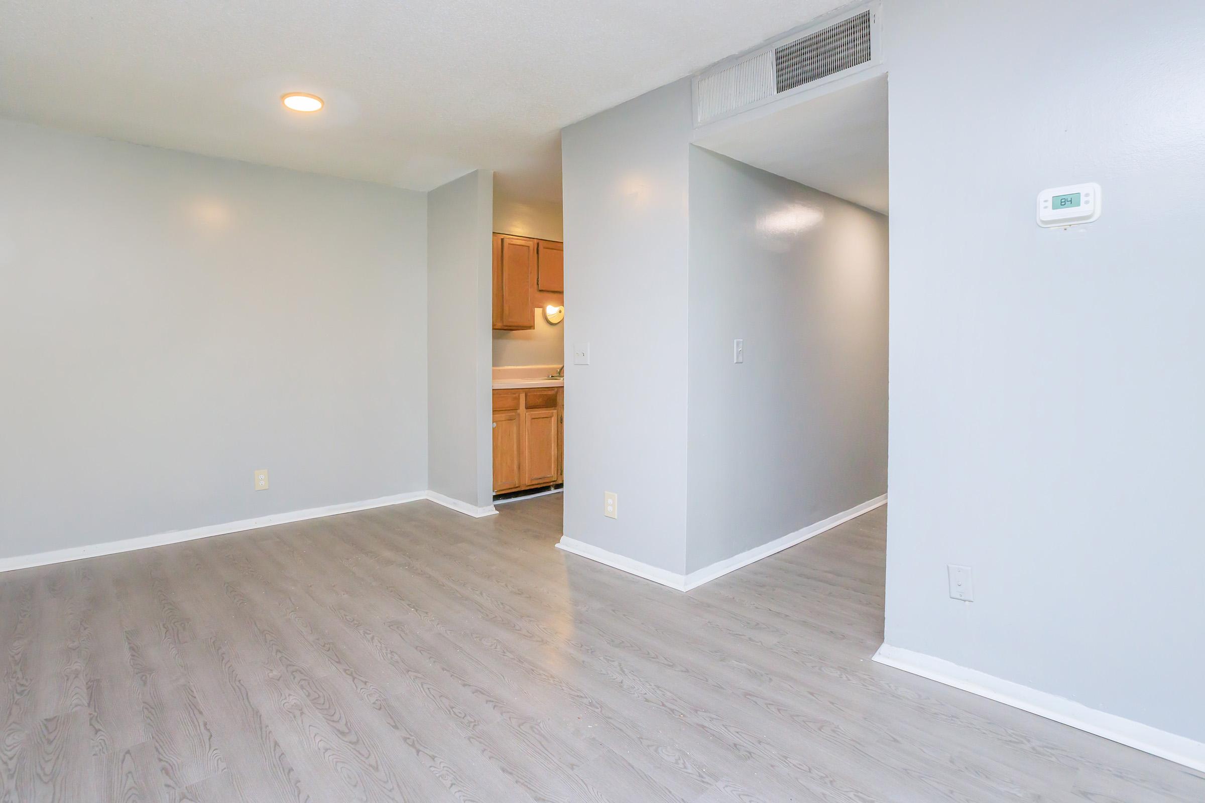 A spacious, empty room with light gray walls and laminate flooring. There is a doorway leading to a kitchen area with wooden cabinets in the background. The room is well-lit with a ceiling light, and there’s a wall thermostat visible to the right.