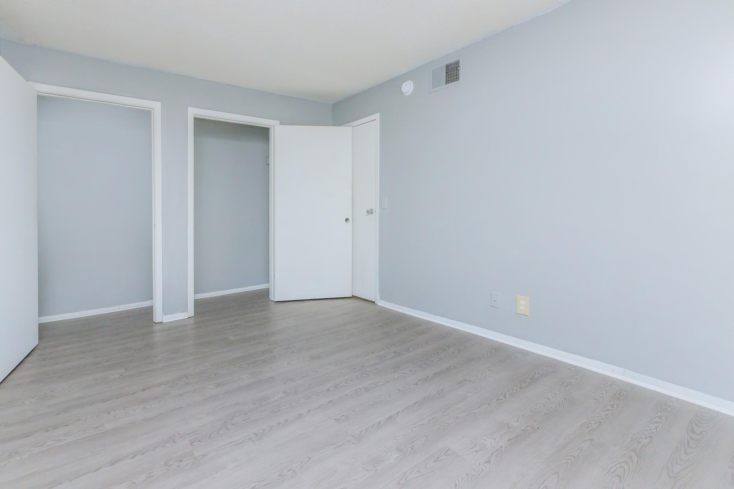 Empty room with light gray walls and light hardwood flooring. There are two doorways leading to other rooms, one of which is partially open. The space is well-lit and minimalistic, suitable for a living area or bedroom. No furniture or decorations present.