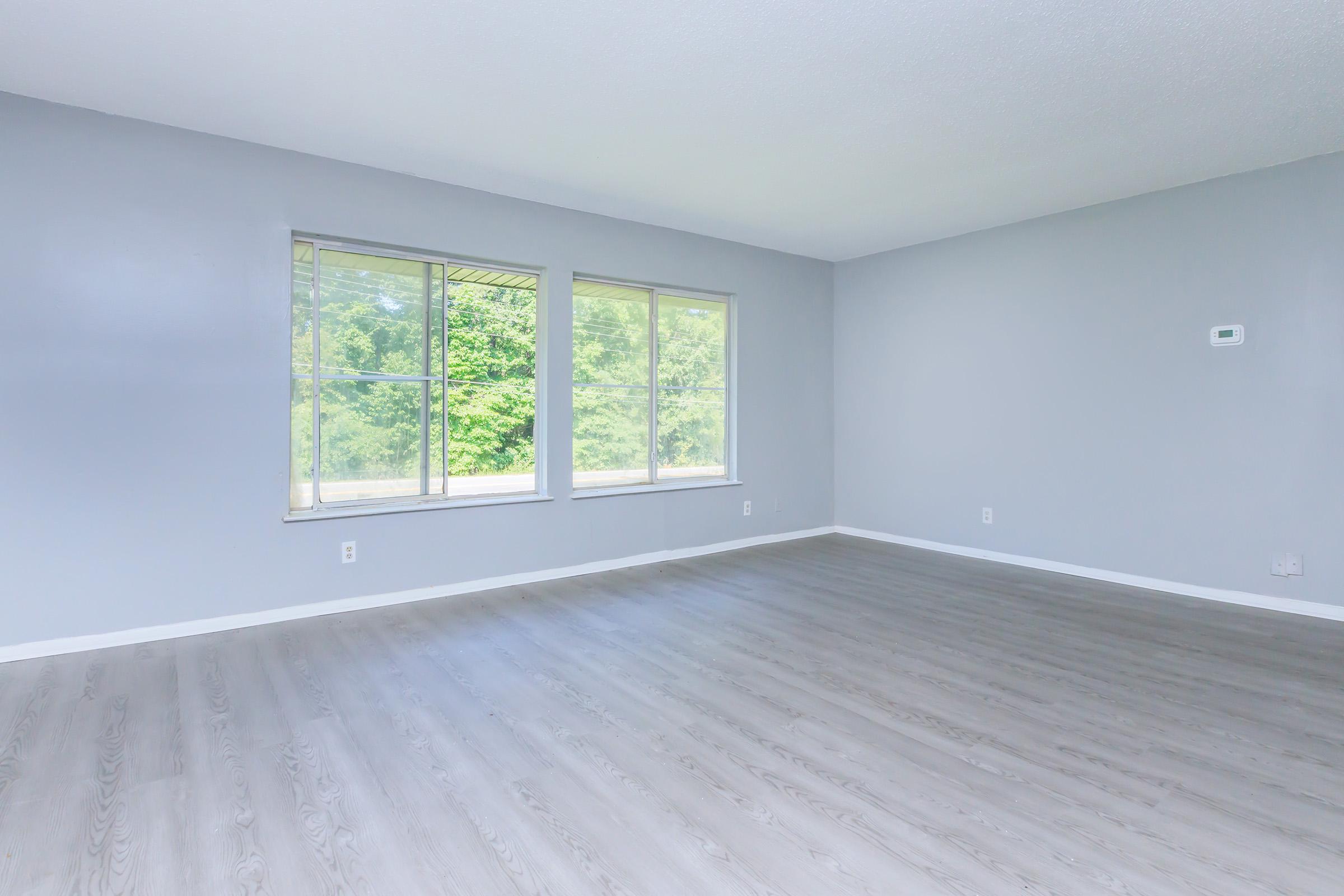 A spacious, empty room with light grey walls and a smooth, light-colored wooden floor. Two large windows let in natural light, showcasing greenery outside. The room has minimal decor and a simple design, ideal for various uses.