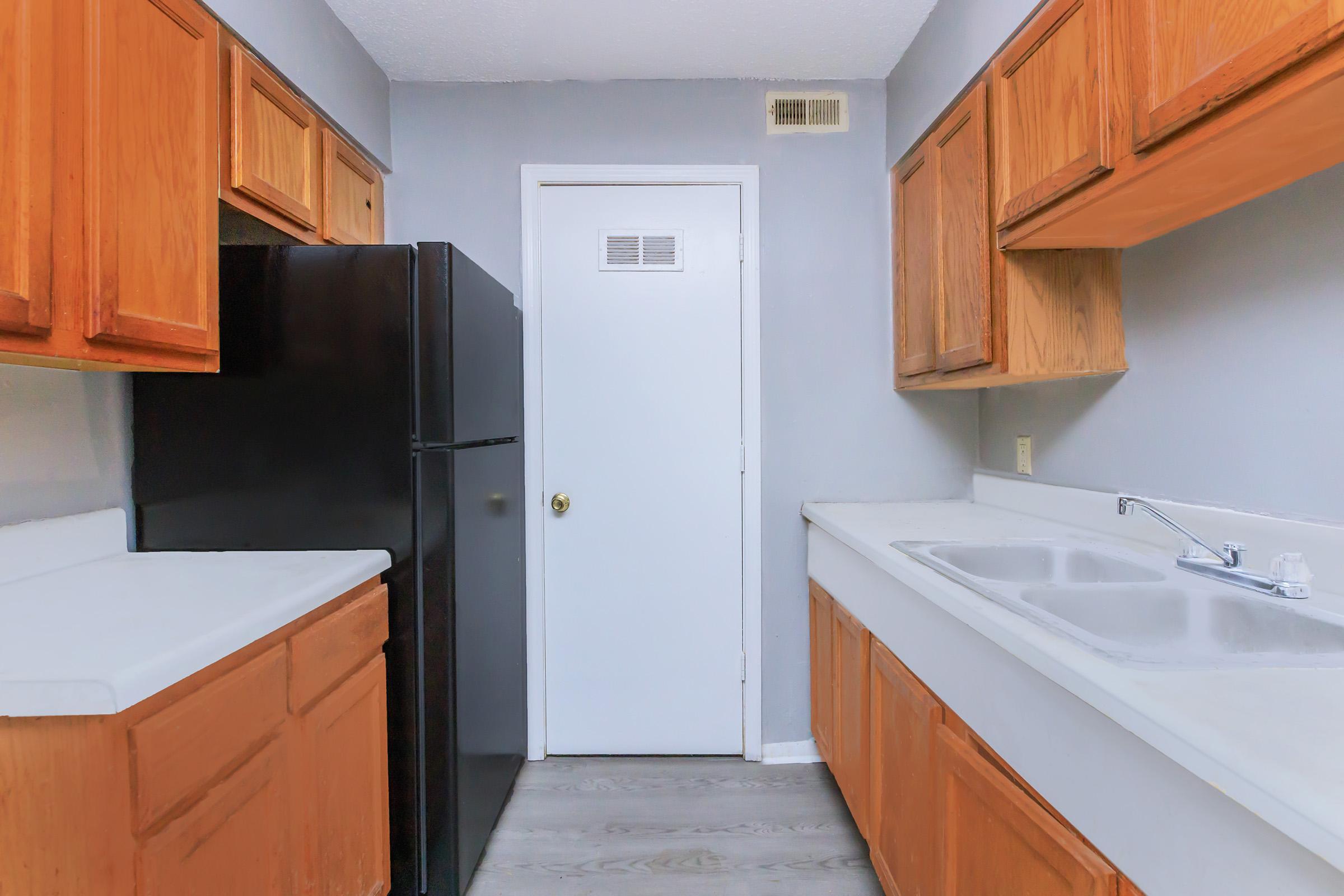 A compact kitchen featuring light-colored walls and wooden cabinetry. The kitchen includes a black refrigerator, a white sink with a faucet, and a countertop. The entrance to another room is indicated by a white door at the end of the kitchen space. The flooring is light-colored, enhancing the overall brightness of the area.