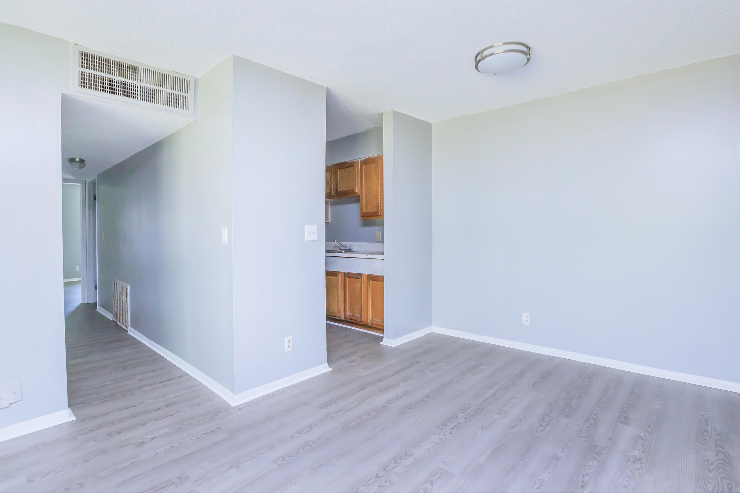 A spacious, light-filled interior of an apartment featuring gray walls, wood-like flooring, and a small kitchen area visible in the background. The layout includes a corridor leading to another room, and overhead lighting fixtures.