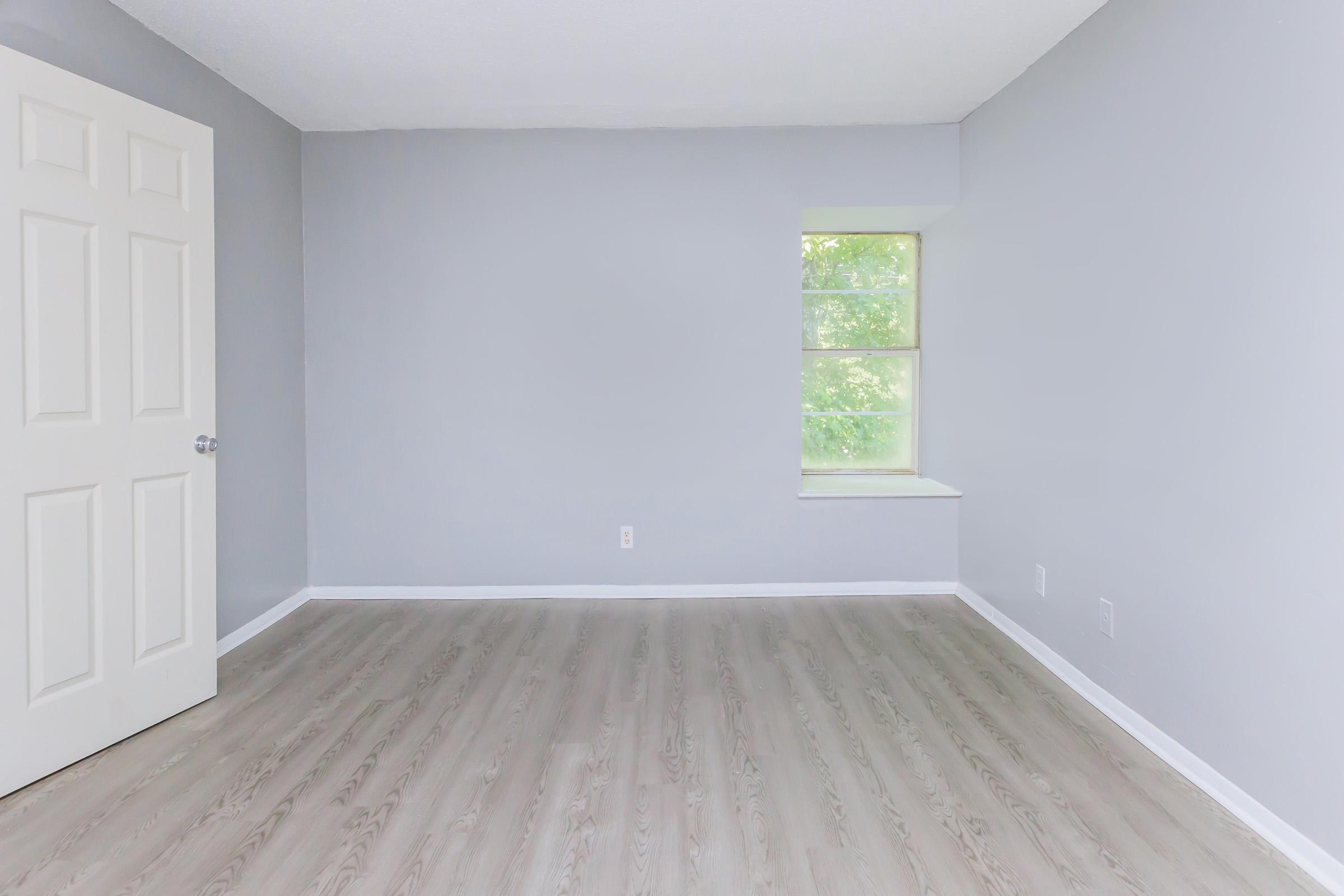 Empty room with light gray walls, a window allowing natural light, and light wooden flooring. A white door is visible on the left side. The room has no furniture, creating a spacious and minimalist appearance.