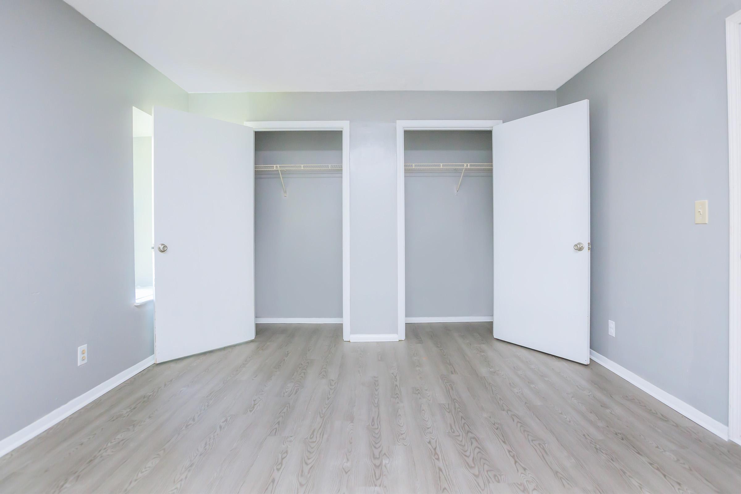 A clean, empty room with gray walls and light wood flooring. Two open closet doors are visible, each with a single shelf inside. Natural light illuminates the space from a window, creating a bright and airy atmosphere.