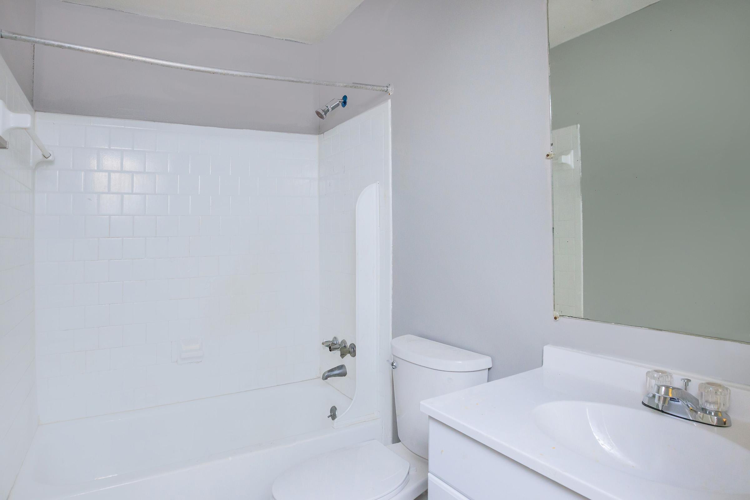 A clean, simple bathroom featuring a white bathtub with a shower, a toilet, and a white sink with a mirror above it. The walls are painted light gray, and the overall design is minimalistic and functional.