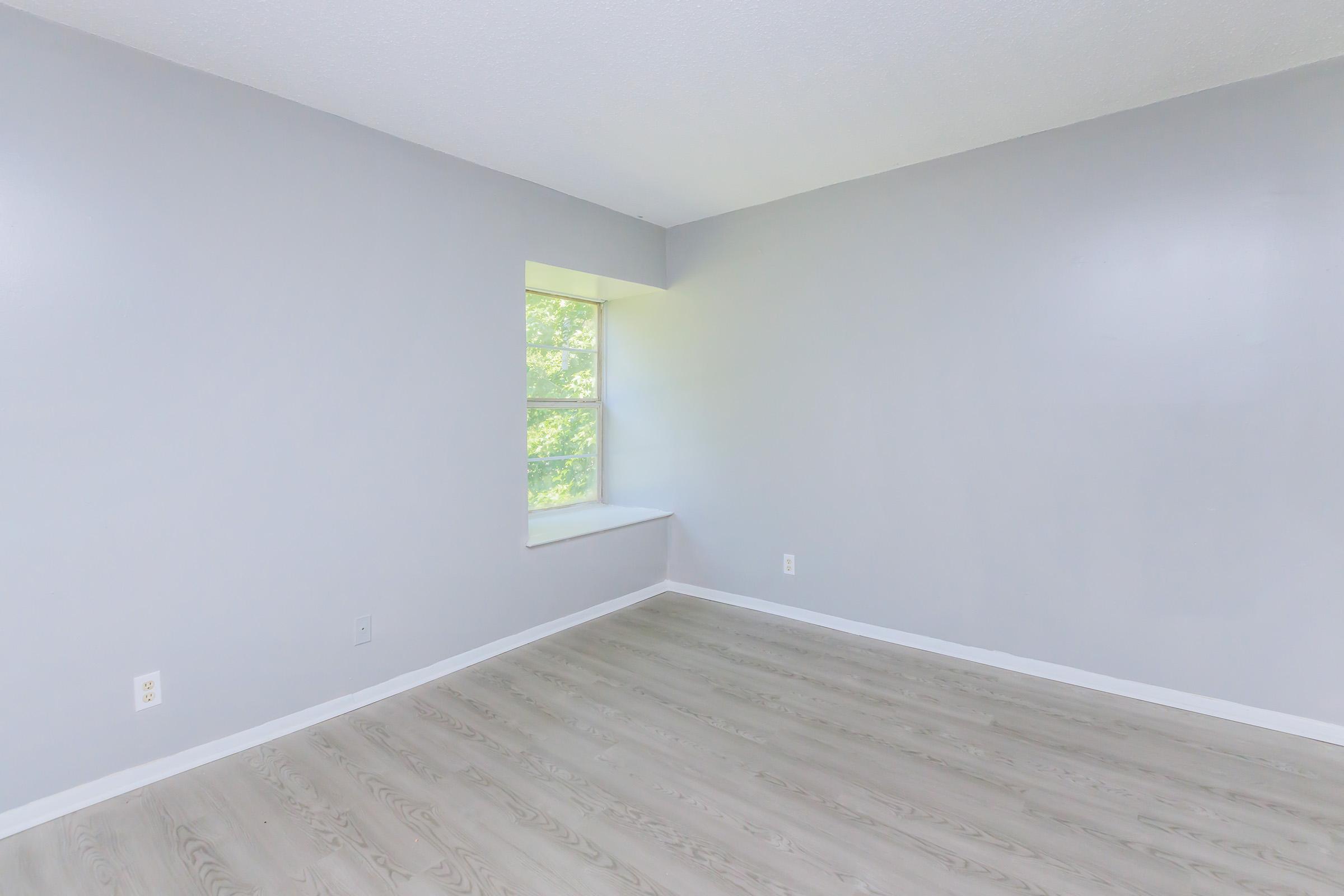 Empty room with gray walls and light-colored wood flooring. A single window with a narrow sill allows natural light to enter, illuminating the space. The room has no furniture or decorations, creating a minimalistic and open atmosphere.