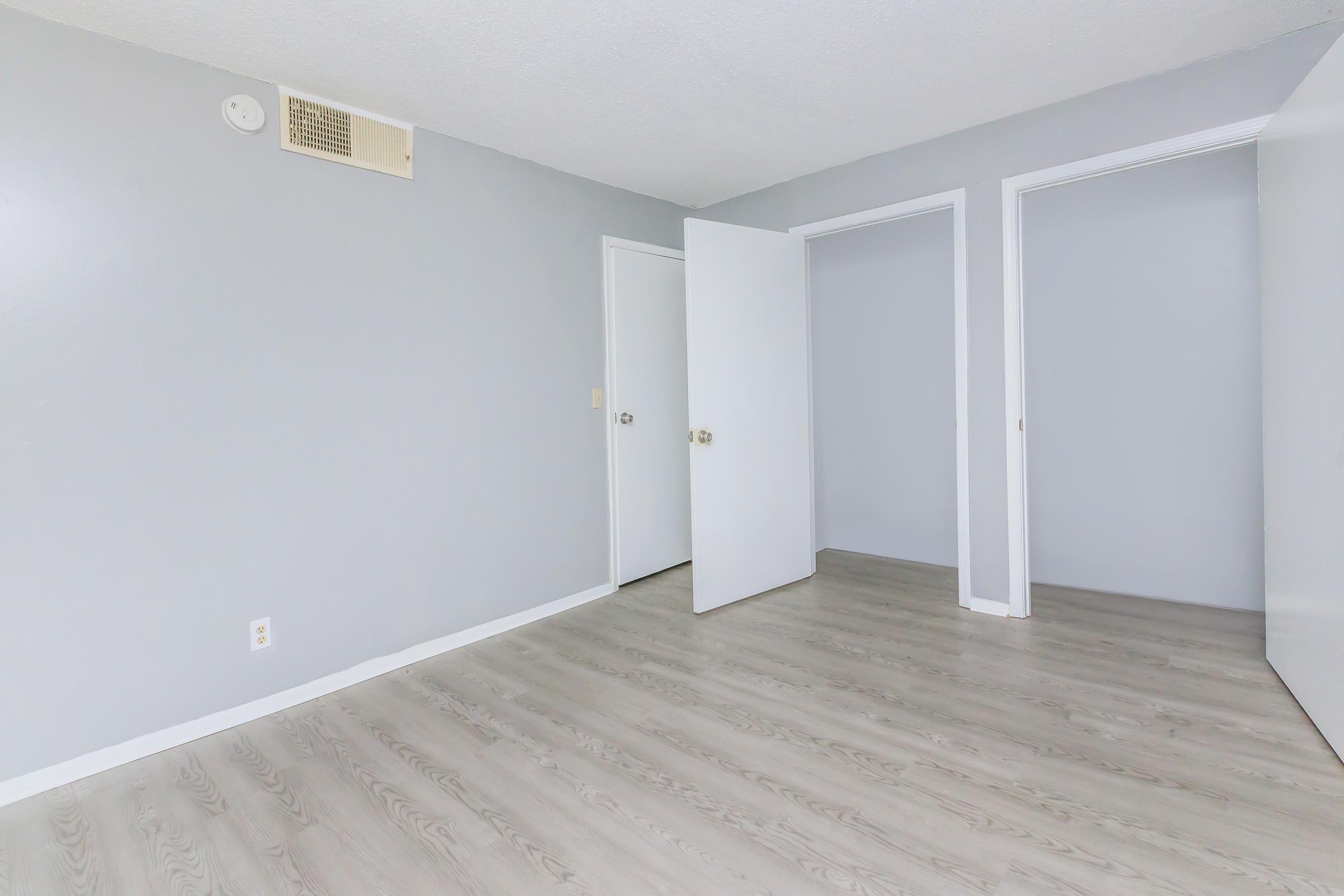 Empty room with light gray walls and laminate flooring. Two doors on the right, one slightly open, leading to separate spaces. The ceiling has a small air vent, and there is a single light source illuminating the area. The overall atmosphere is clean and minimalistic.