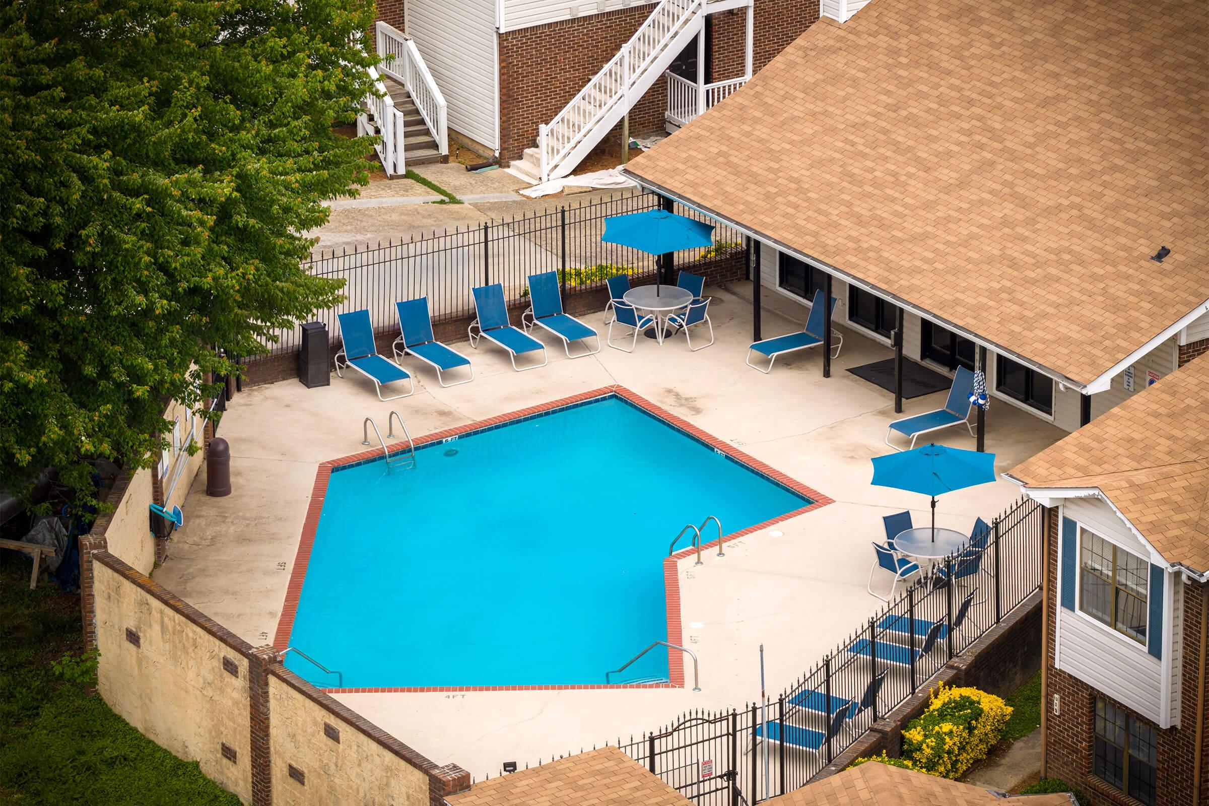 Aerial view of a swimming pool area featuring an octagonal blue pool, surrounded by lounge chairs and umbrellas. The pool is enclosed by a black fence, with a building visible in the background and greenery nearby.