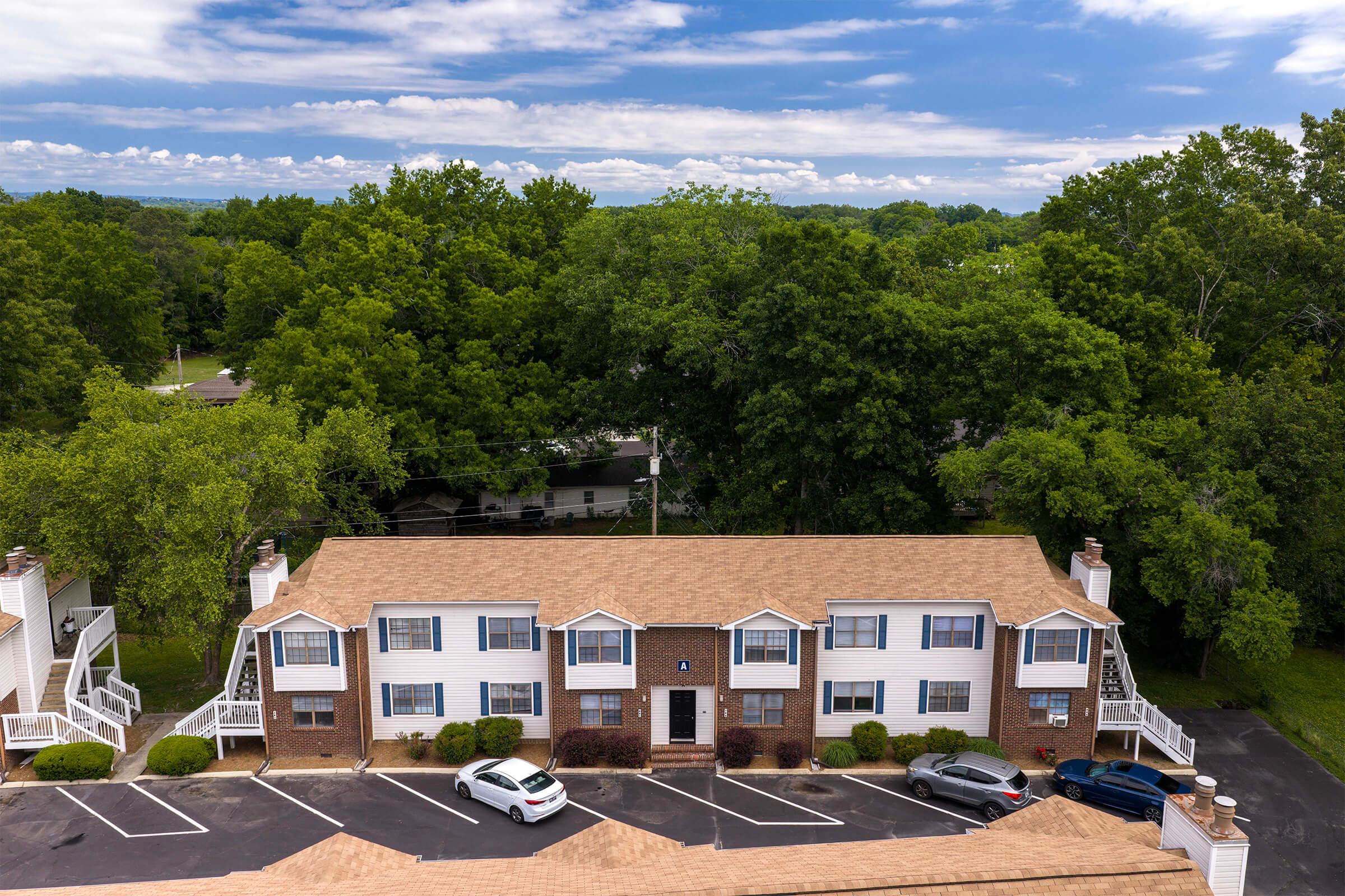 Aerial view of a multi-unit apartment building with a tan asphalt roof, surrounded by lush green trees. The building features several windows and white siding. A parking lot is visible in front with parked cars, and the sky is partly cloudy, adding a serene atmosphere to the scene.