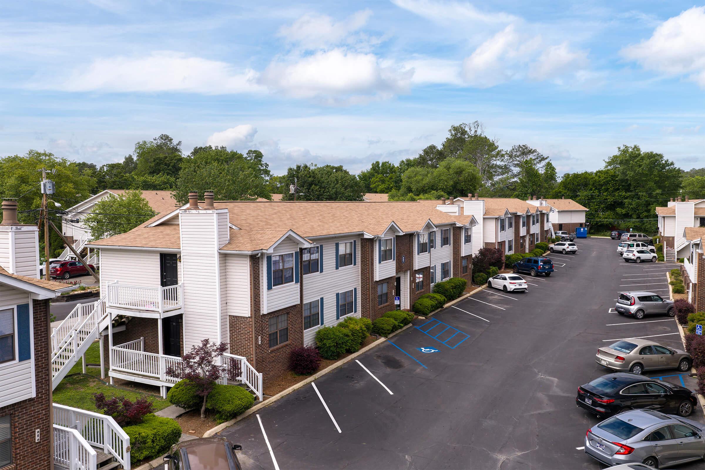 A view of a residential apartment complex featuring two-story buildings with white siding and brick accents. Several parked cars are along the blacktop parking lot, surrounded by landscaped greenery. The sky is partly cloudy, indicating a bright day.