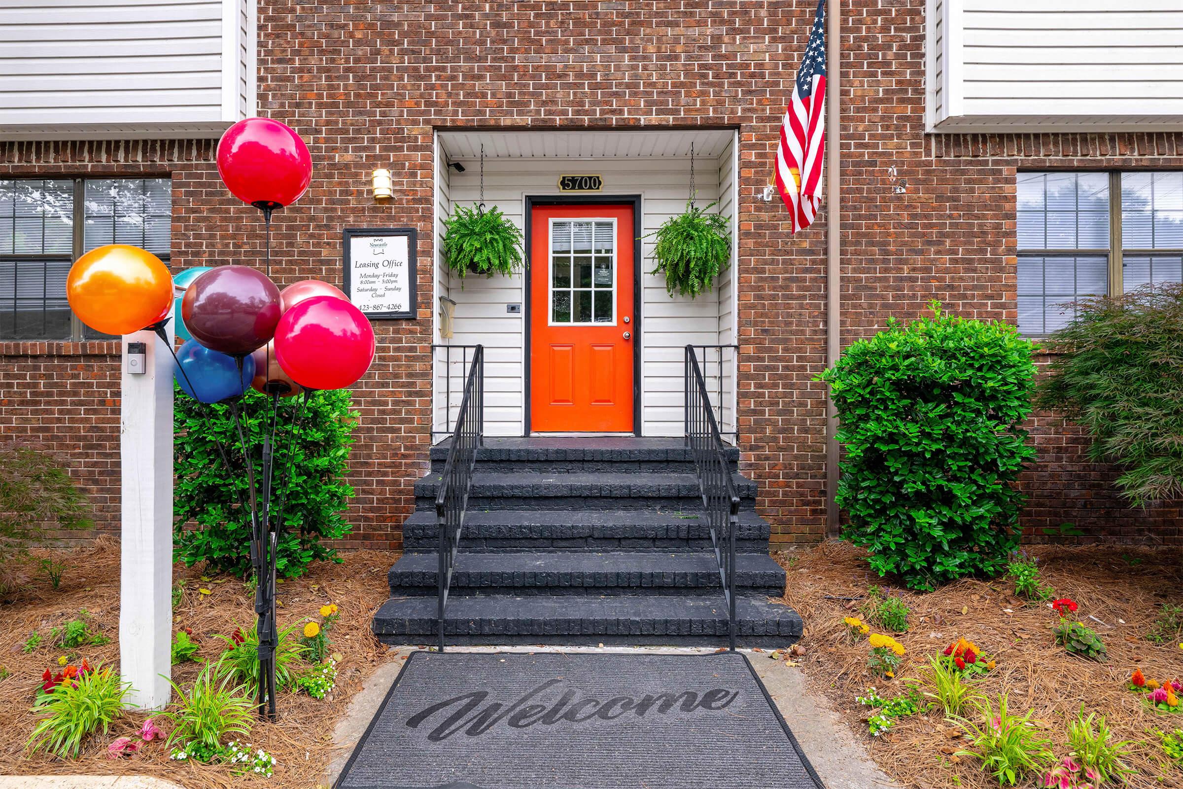 Front entrance of a building featuring a bright orange door, welcome mat, and stairs leading up. On the left, colorful balloons are tied to a railing, and there are green plants in hanging pots. An American flag is displayed on the side of the building, adding a festive touch to the welcoming atmosphere.