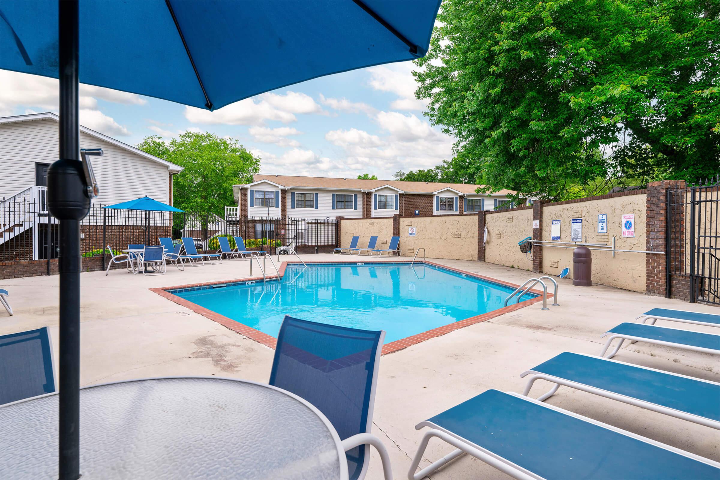 A serene outdoor pool area featuring a clear blue pool surrounded by lounge chairs. An umbrella provides shade over a nearby table. The area is bordered by a fence, with residential buildings and trees visible in the background against a partly cloudy sky.