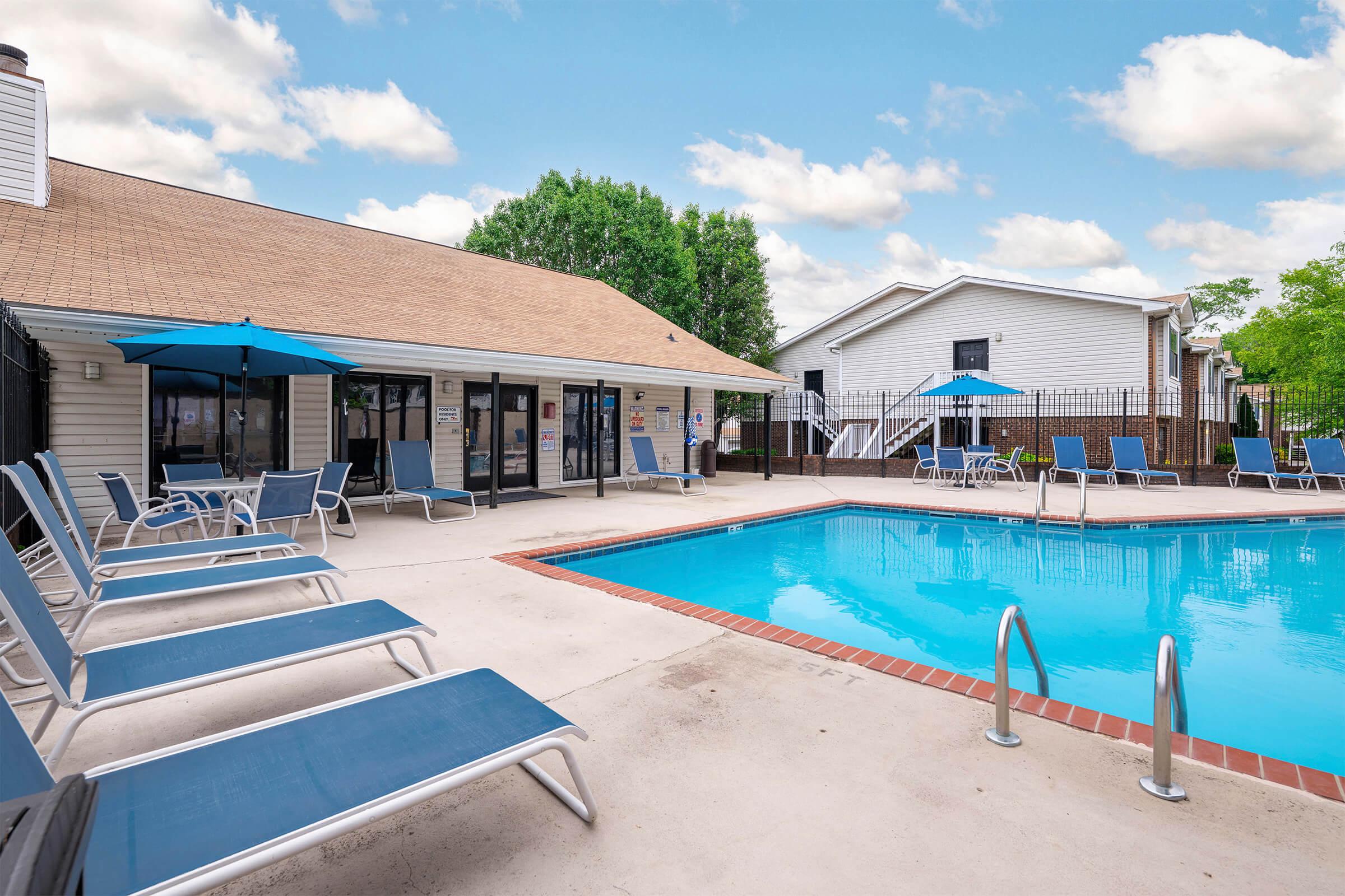 A sunny outdoor pool area featuring a blue swimming pool surrounded by lounge chairs, with a clubhouse in the background. Parasol umbrellas provide shade, and the area is enclosed by fencing, creating a relaxing atmosphere for leisure and enjoyment. The sky is partly cloudy.