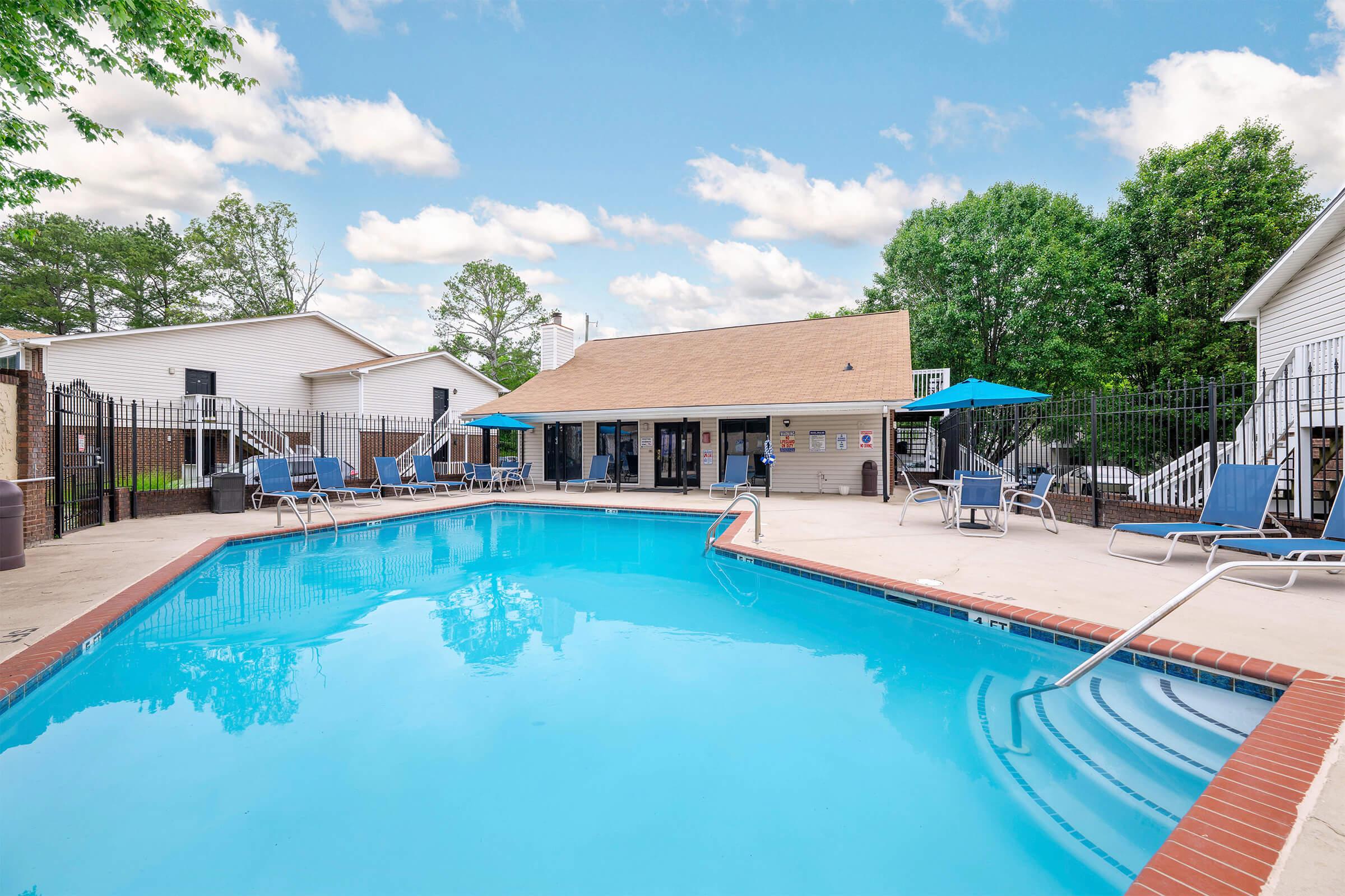 A clear blue swimming pool surrounded by lounge chairs and umbrellas. In the background, a building with a tan roof is visible, along with green trees and a bright blue sky with white clouds. The area is fenced, creating a cozy outdoor space for relaxation.