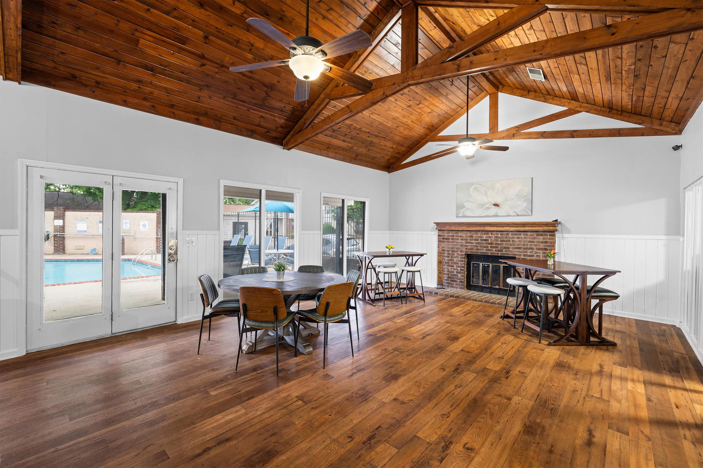 A spacious indoor area with wooden beams and flooring, featuring tables and chairs arranged neatly. Two ceiling fans are visible, and large glass doors lead to an outdoor pool area. A cozy fireplace and a floral artwork adorn the wall, creating a welcoming atmosphere for gatherings or events.