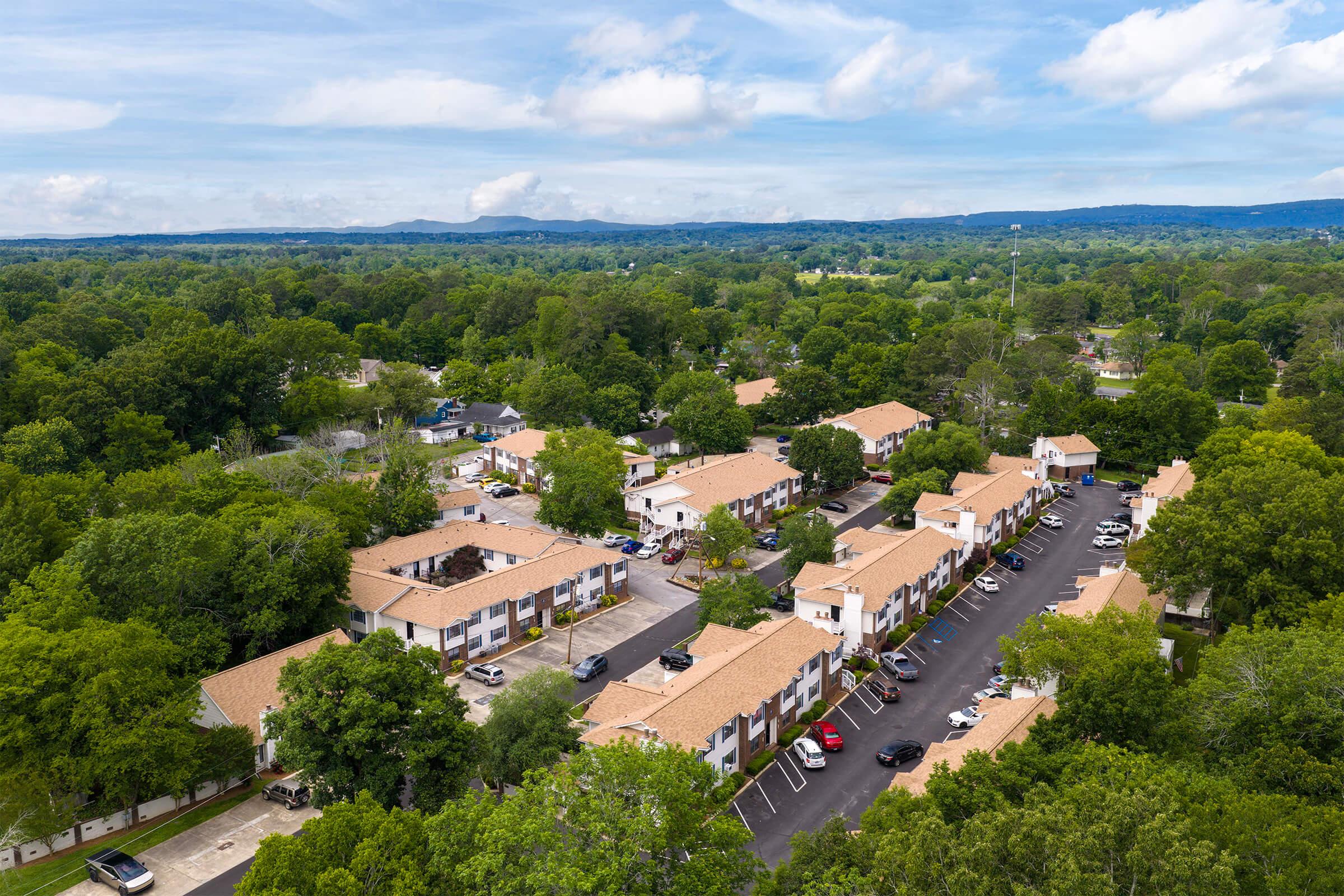 An aerial view of a residential area featuring multiple two-story buildings with red roofs surrounded by lush green trees. The scene includes parked cars along a winding road, with distant hills visible under a partly cloudy sky.