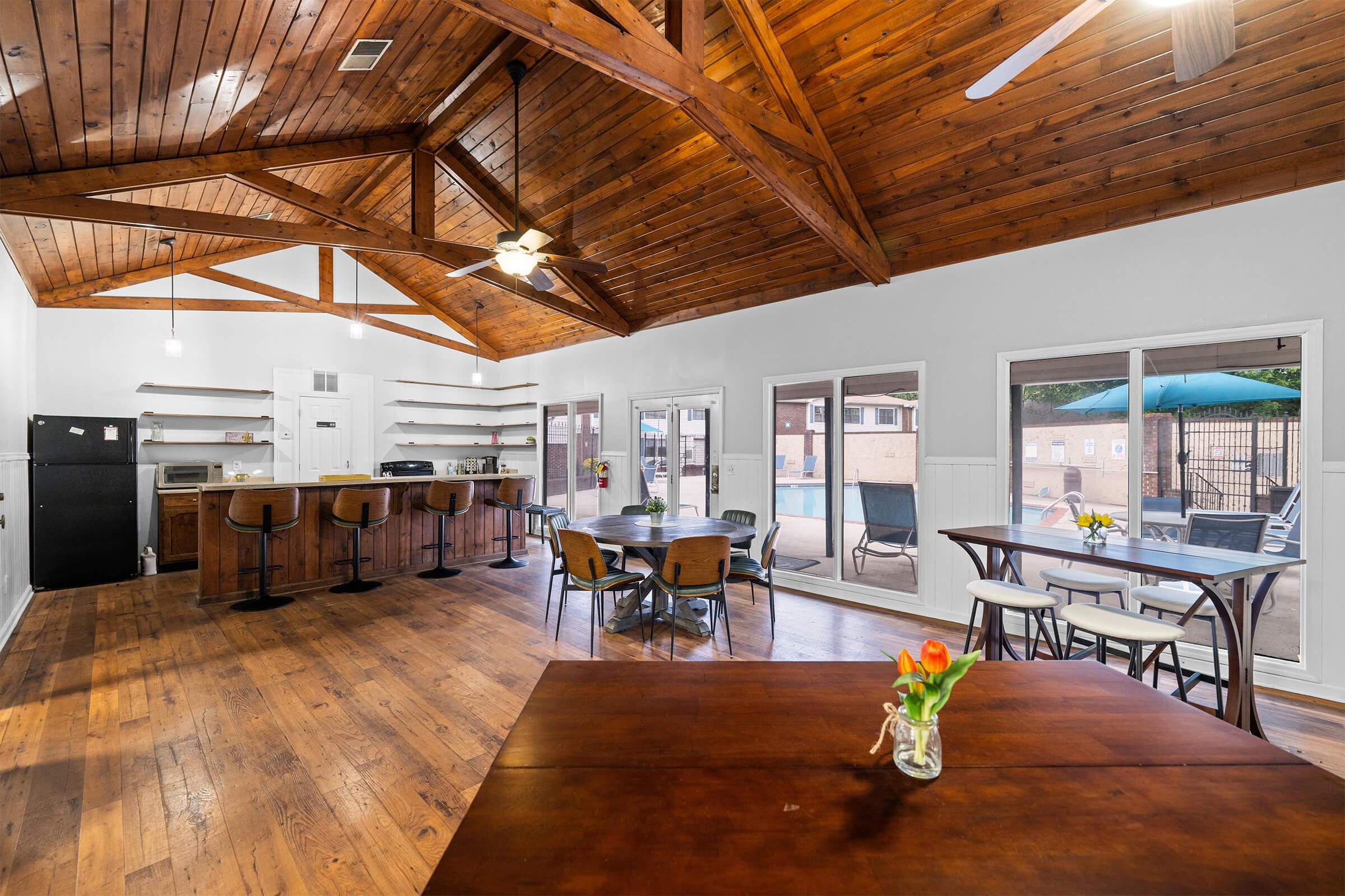 Spacious modern kitchen and dining area with a high wooden ceiling. Features include a large dining table with a vase of flowers, bar seating, and large windows that provide natural light. The room opens up to a patio area with outdoor seating.